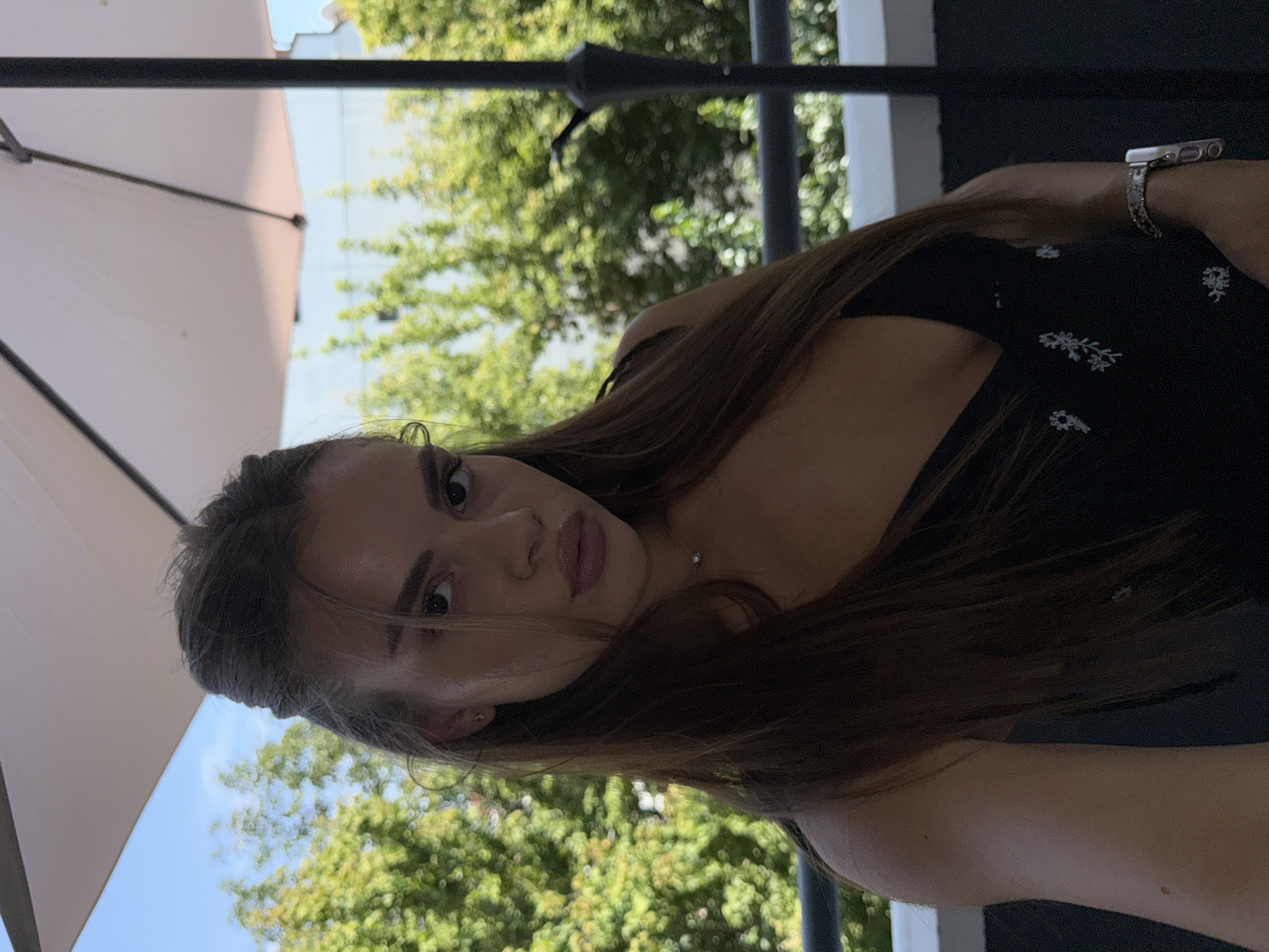 A young woman with long, brown hair and a black dress is pictured outdoors under a large white umbrella. She is wearing a silver necklace and a watch on her left wrist. In the background, green trees and a blue sky are visible, suggesting a sunny day. The scene appears to be on a balcony or terrace, possibly in Plovdiv, Bulgaria, given the architecture and foliage which are typical of the region.