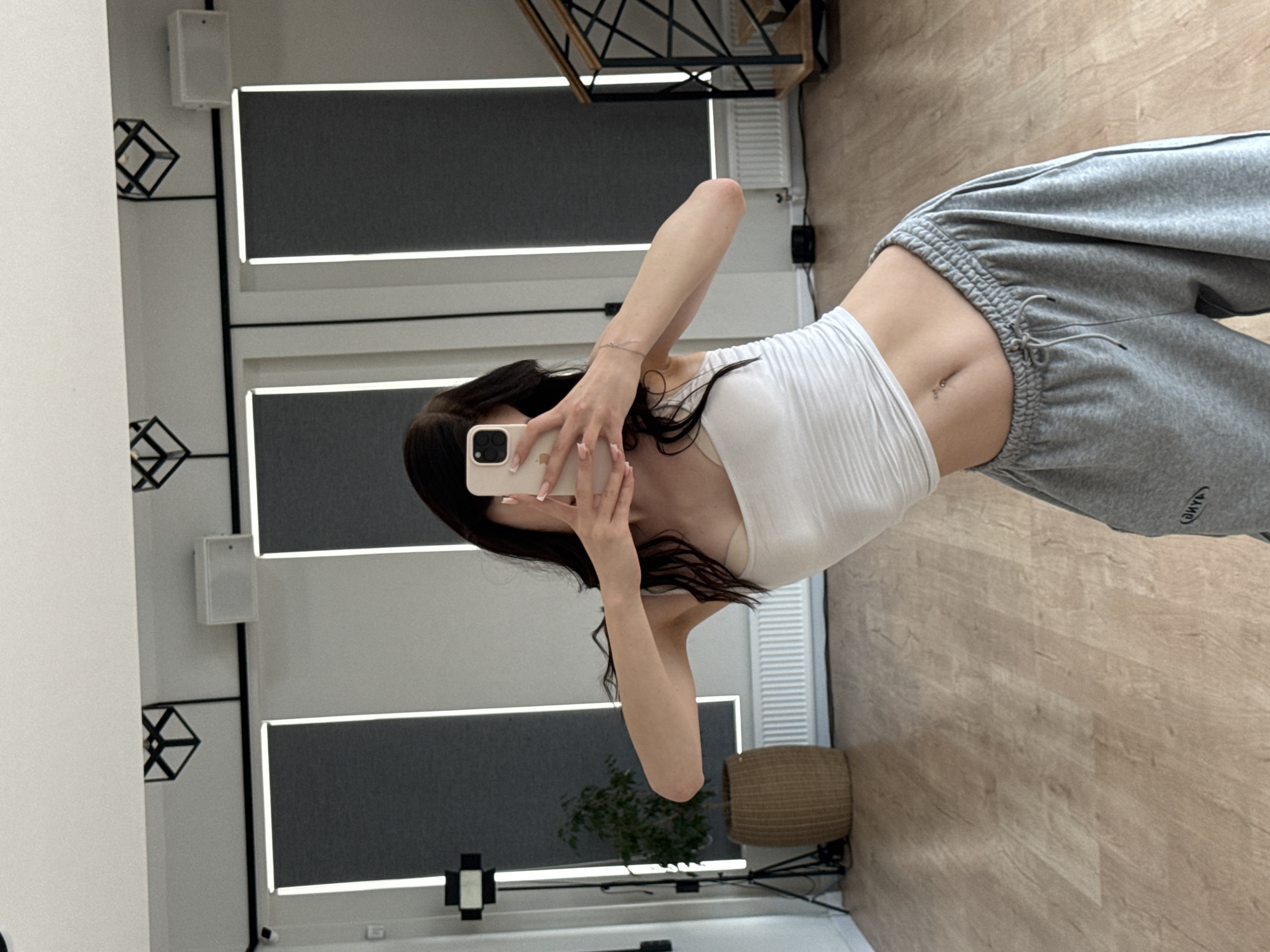 The image shows a young woman taking a selfie in a well-lit room with a light wood floor. She is wearing a white strapless top and grey sweatpants. She has long dark hair and a belly button piercing. The room appears to be a modern space, possibly a home or studio, with sleek minimalist decor. In the background, there are large rectangular panels with thin white light strips, and on the left wall, there are cubic decorative shelves and a white speaker. A woven basket and a plant are visible in the corner of the room. The overall impression is one of casual relaxation and personal style. The lighting suggests it is daytime. There is visible text on the sweatpants which appears to read "CYBER".