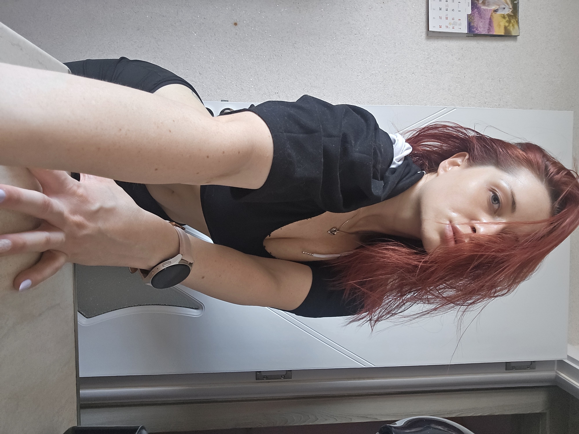 The image displays a portrait of a person, likely female, taken from an elevated angle and rotated 90 degrees counter-clockwise. The subject's upper body is visible, with their head resting on a bent left arm, which is positioned on a light-colored surface in the foreground. They gaze directly at the camera with a calm, slightly parted-lips expression.
The person has long, vibrant red or auburn hair, appearing somewhat disheveled and falling across their face and chest. They are wearing a black V-neck top, possibly a crop top, revealing a small part of their midriff and décolletage. A delicate necklace with a small pendant adorns their neck. On their left wrist, a light-colored smartwatch with a rose gold or pink band is clearly visible. Their fingernails are painted with a subtle lavender or periwinkle polish, and their skin shows a scattering of freckles.
The background suggests an indoor setting. A light grey wall is visible, on which a calendar for the year "2020" hangs, featuring a white horse in a lavender field. Below the calendar, a white door or cabinet is slightly ajar. The immediate foreground consists of a light-toned countertop. The scene conveys a relaxed and intimate moment. The location context for the image is Burgas, Bulgaria.