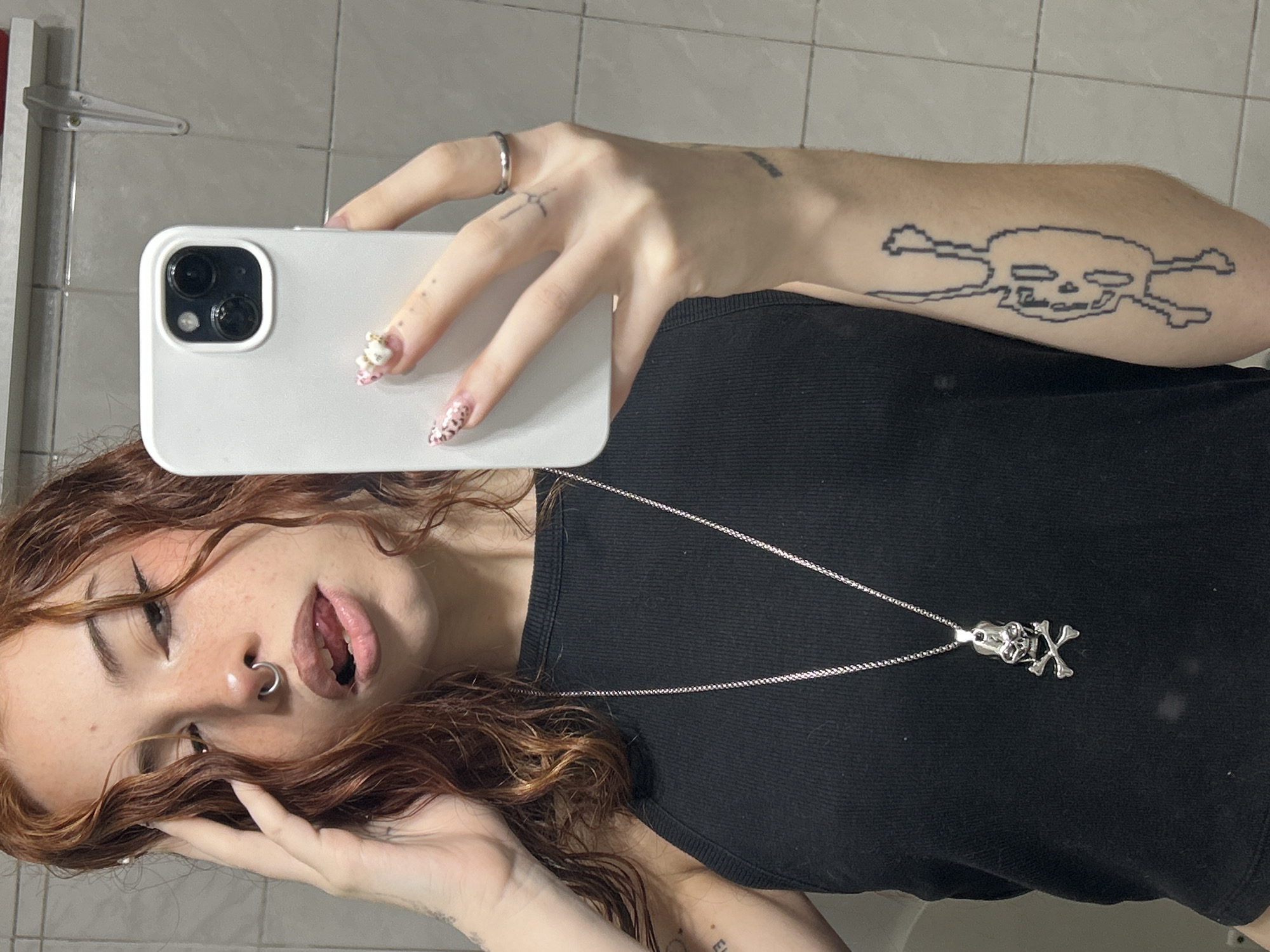 A young woman with red hair and a nose ring is taking a selfie in a bathroom. She is holding a white smartphone in her left hand, and her right hand is near her face. She is wearing a black tank top and a silver necklace with a skull and crossbones pendant. The woman has tattoos on her arms. The background shows a tiled wall, suggesting a bathroom setting. The location is unknown.