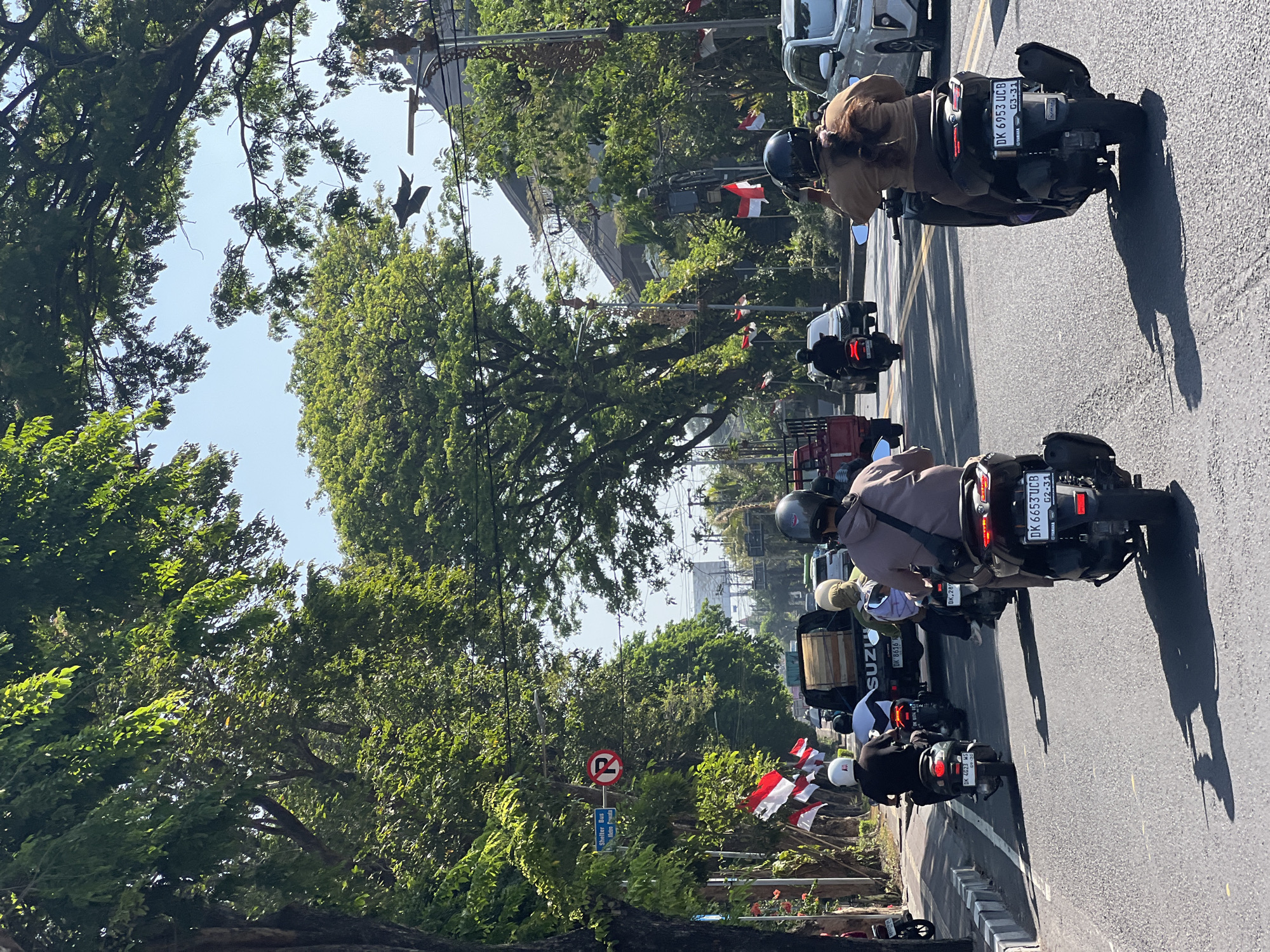 The image depicts a sunny day in Denpasar, Indonesia, with several motorbikes navigating a tree-lined street. The road is paved, with yellow and white lane markings visible. Indonesian flags are prominently displayed along the roadside, suggesting a festive or patriotic occasion. Trees with lush green foliage overhang the street, casting shadows on the asphalt. In the background, buildings and power lines are visible. The focus is on the motorbikes and their riders, who are all wearing helmets. Some riders are looking ahead, while others appear to be interacting with each other or their surroundings. One motorbike in the foreground displays a license plate with the characters "DK 6953 UCB." The overall atmosphere is one of bustling urban activity on a bright, clear day.