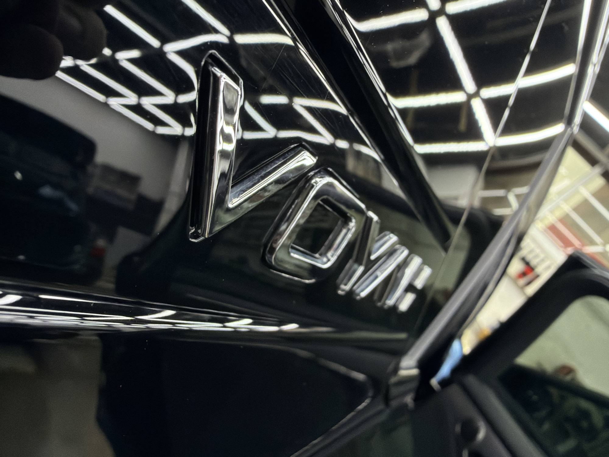 A close-up, eye-level shot focuses on a glossy black surface, likely a car's body panel, featuring a prominent chrome emblem that spells "VOYAH" in stylized capital letters. The highly reflective surface captures a clear, grid-like pattern of bright overhead lighting, characteristic of a controlled indoor environment such as a car showroom, detailing bay, or workshop. Numerous fine scratches are visible on the black paintwork, particularly around and on the emblem itself, indicating wear or imperfections. The chrome letters of the "VOYAH" emblem also show reflections and slight variations in their polished finish.
The background, largely out of focus, further emphasizes the indoor setting with more distorted reflections of the same grid lighting and indistinct shapes that could be parts of a vehicle's interior or adjacent objects. No people or specific activities are visible, but the detailed view of the emblem and the visible scratches suggest a moment of inspection or presentation of the vehicle. The lighting is artificial and bright, allowing for clear observation of surface details. No specific time of day or weather can be inferred. The image captures the aesthetic of a premium vehicle's branding under intense scrutiny.