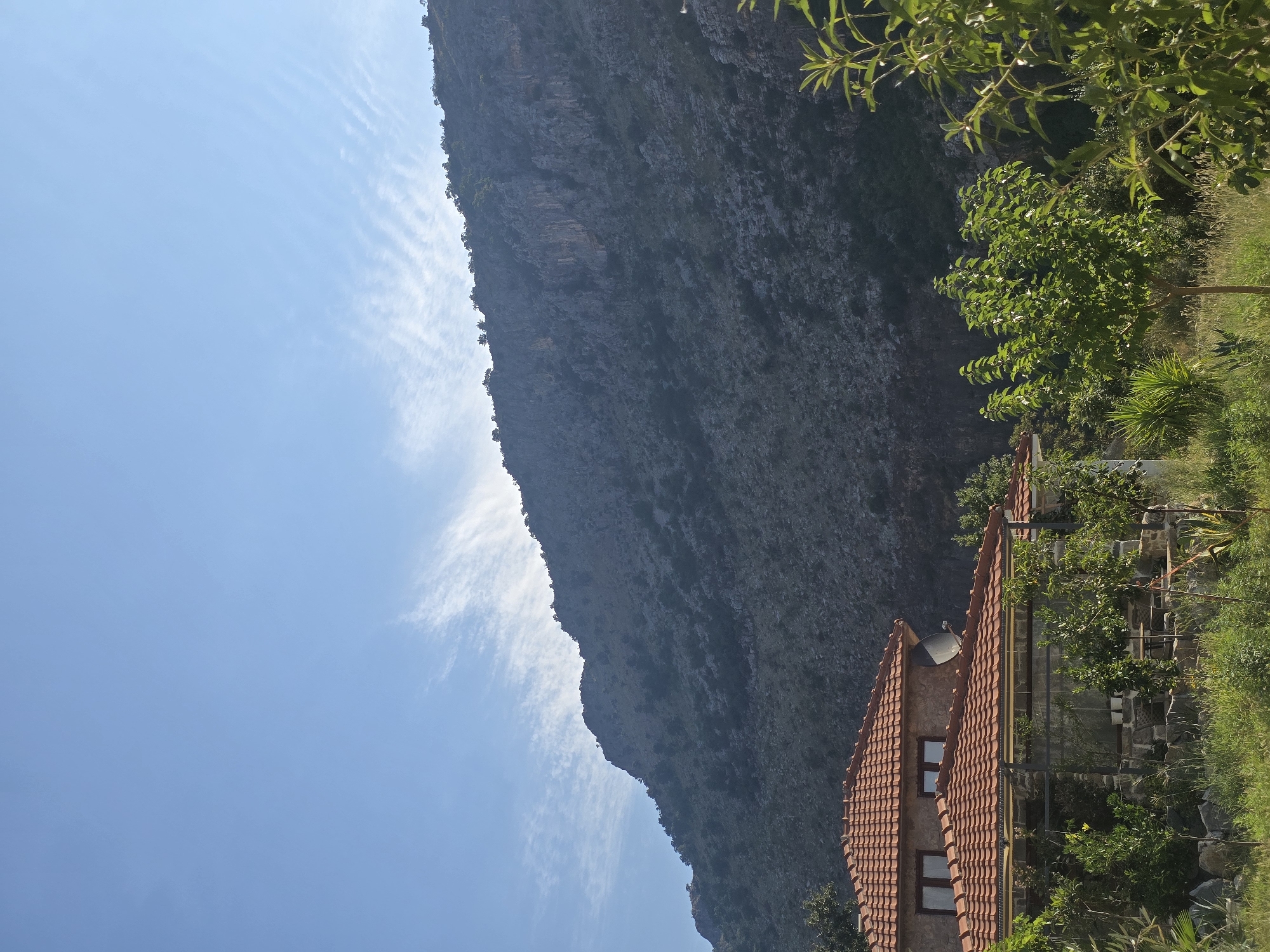 The image depicts a serene landscape featuring a house with a red-tiled roof nestled in a lush green area. In the background, a majestic mountain rises under a clear blue sky adorned with wispy clouds. The foreground is dominated by dense vegetation, including trees and grass, creating a natural and inviting setting. The house appears to be a single-family dwelling, constructed with stone and featuring windows. A satellite dish is visible on the roof, suggesting modern amenities. The overall scene evokes a sense of tranquility and rural charm, possibly located in a suburban or mountainous region near Podgorica, Montenegro, given the geological features of the landscape. No people are visible in the image.