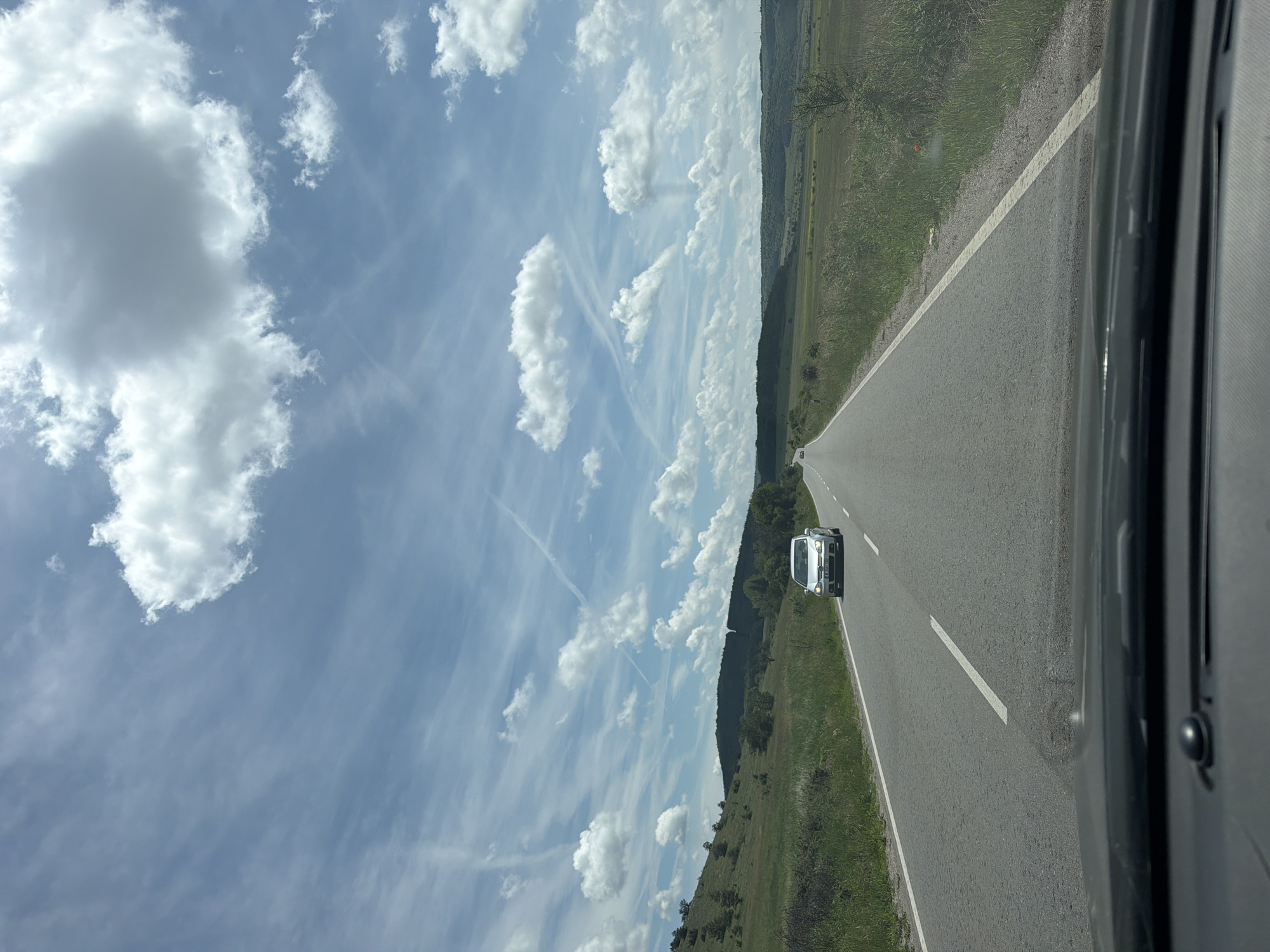 The image, correctly oriented horizontally, presents a view from inside a vehicle traveling on a two-lane asphalt road through a rural landscape. The road stretches into the distance, with a light-colored car, possibly silver, visible ahead on the same side of the road. The car's headlights are on.
To the left of the road, there is a wide expanse of green grass or a gently sloping hillside, leading up to a distant, tree-covered mountain range or large hills. The right side of the frame is occupied by the dark interior trim of the viewer's vehicle, likely a window frame or door, with a narrow grassy verge visible beside the road. A small red object is barely discernible on the grassy shoulder to the far right.
The sky is a prominent feature, displaying a bright blue color adorned with numerous white, puffy cumulus clouds and thinner, streaky cirrus clouds or contrails. The overall weather appears sunny and clear.
Notable details include the white solid line delineating the right edge of the road. The license plate of the car ahead clearly shows "CB," which is a registration code for Sofia, Bulgaria, confirming the general location provided (Zavidovtsi, Bulgaria). The scene conveys a sense of open-road travel through a picturesque, hilly countryside during the daytime.