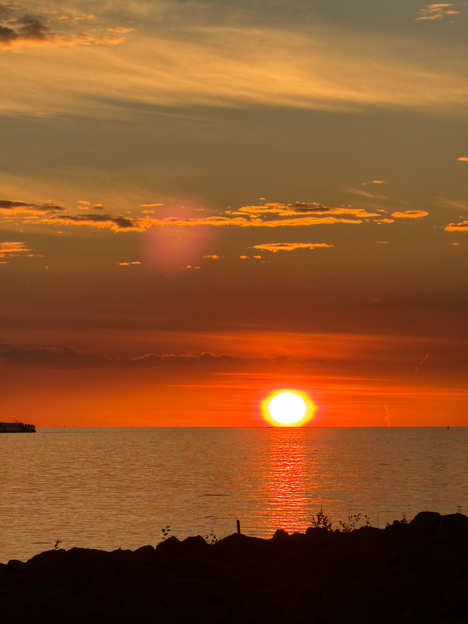 A vibrant sunset over the Gulf of Finland, as viewed from the coastline of Saint Petersburg, Russia. The sun, a brilliant white orb with a golden halo, is positioned near the horizon, casting a fiery orange glow across the sky and water. Wispy clouds, tinged with orange and yellow, are scattered throughout the upper atmosphere. The water's surface reflects the sunset's colors, creating a shimmering path of light leading towards the viewer. In the foreground, a silhouetted rocky shoreline with sparse vegetation provides a dark contrast to the luminous sky and sea. On the left side of the image, a distant, dark structure, possibly a pier or vessel, is visible near the waterline. The overall scene is serene and picturesque, capturing the dramatic beauty of a seaside sunset.