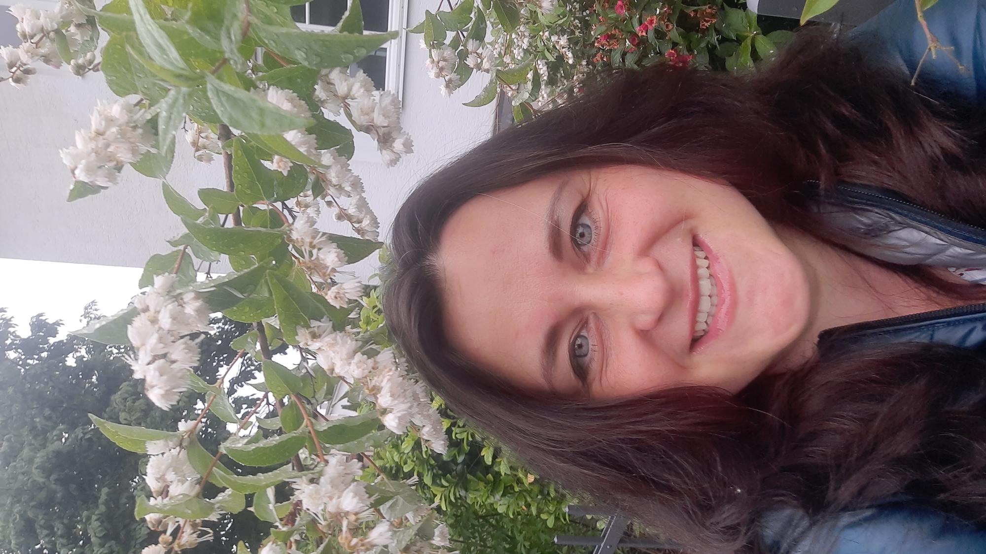 The image is a close-up, selfie-style photograph of a woman with long, dark brown hair and blue eyes, smiling broadly at the camera. She is wearing a dark blue jacket with a silver lining. Behind her, there are branches of a flowering bush with small white blossoms and green leaves, some of which appear to have water droplets on them. A white building with a window is visible in the background, as is some greenery. The scene suggests an outdoor setting, possibly a garden or residential area. The image does not provide specific details about the location within the city of Rosenheim, Germany.