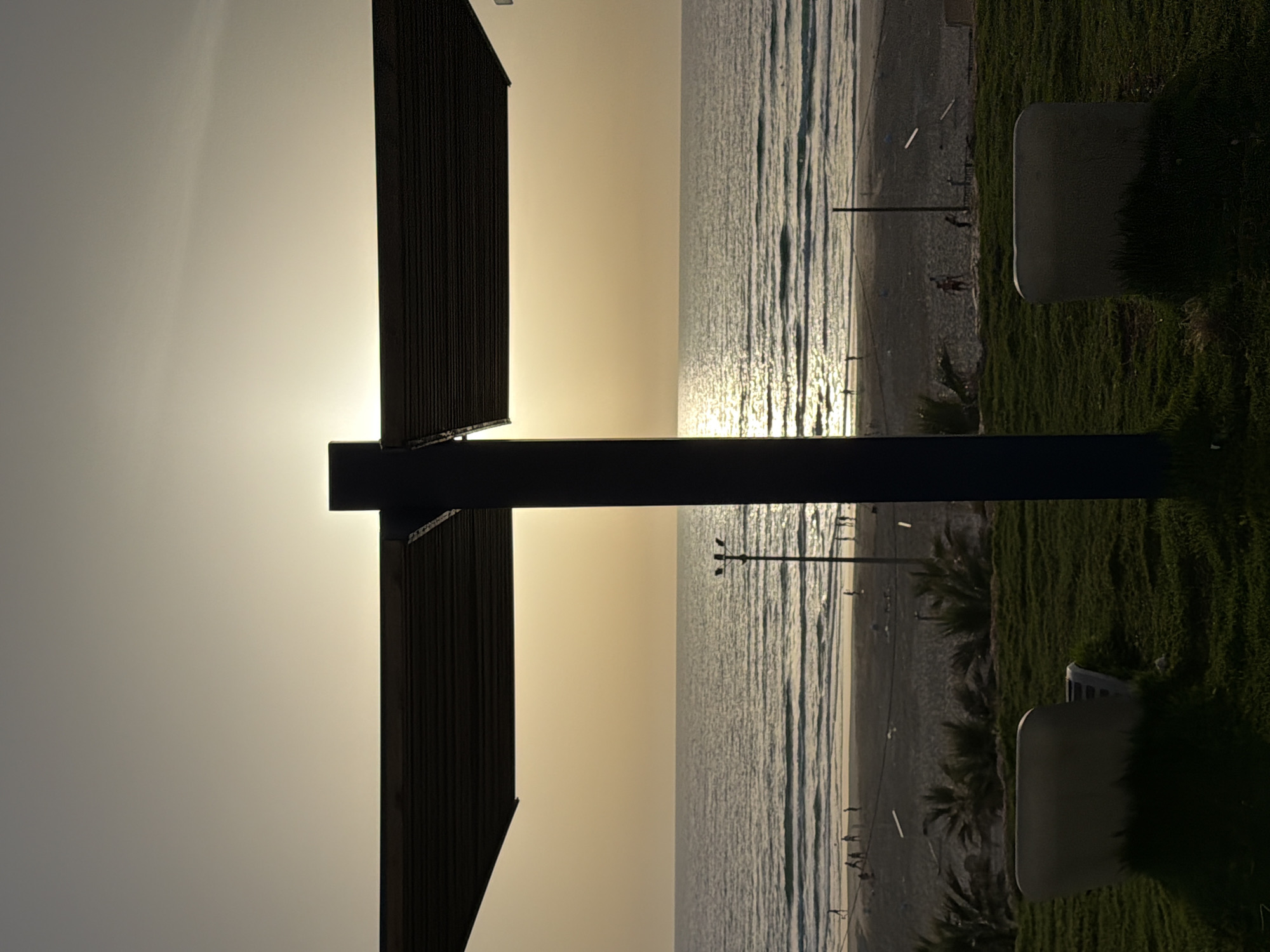 The image shows a seaside scene in Rishon LeZion, Israel. The view is framed by a dark, vertical architectural structure on the left, possibly part of a building or a decorative installation. The bright sky and the sun, creating a golden glow, are visible between the structure and the sea.
The sea itself occupies a significant portion of the image, with visible wave patterns and sunlight reflecting on the water's surface. The coastline is lined with a sandy beach, dotted with people, some walking along the shore.
On the right side of the image is a grassy area. Two light-colored, possibly concrete benches are present in this green space. Palm trees are scattered along the coastline, suggesting a warm, coastal climate. The presence of electrical poles along the beach indicates the area is likely a public space.