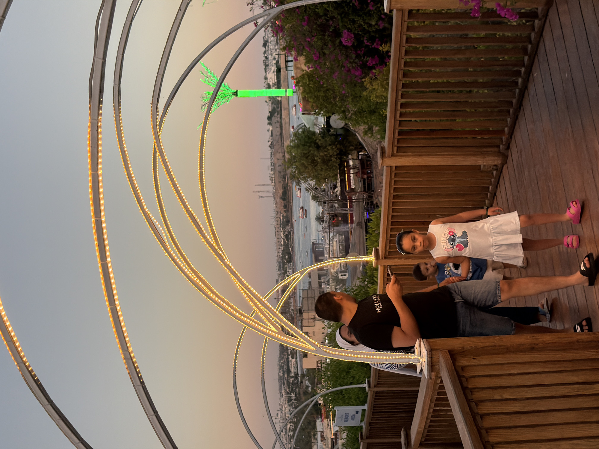 In Sharm El Sheikh, Egypt, an image captures a moment on a wooden boardwalk with arched, illuminated structures overhead. The boardwalk is lined with wooden railings. Several people are visible. In the foreground, a young girl in a white dress with a Stitch graphic and pink sandals is walking towards the viewer. Behind her, a young boy in a blue t-shirt and shorts is also walking. To the left, a man in a black t-shirt and denim shorts is looking at his phone, with a woman in a white headscarf partially visible behind him. Further in the background, a cityscape with buildings and a body of water with boats can be seen. A prominent, brightly lit green palm tree sculpture stands out against the twilight sky, which transitions from a pale blue to an orange hue. The overall scene suggests an evening setting, possibly at a resort or entertainment area in Sharm El Sheikh.