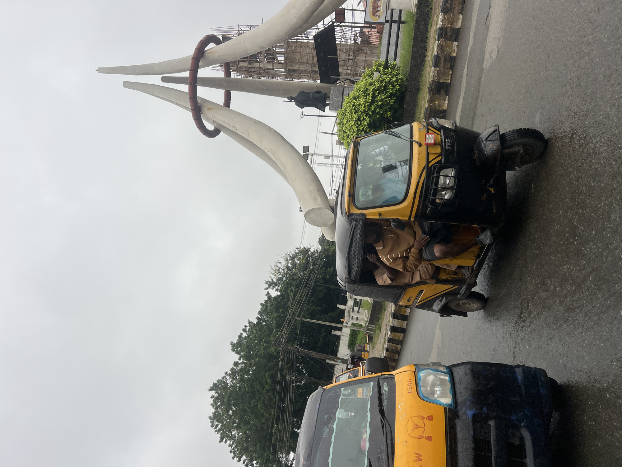 The image depicts a street scene in Onitsha, Nigeria, under an overcast sky. In the foreground, a yellow and black auto-rickshaw, known locally as a Keke Napep, is captured in motion. Three male passengers are visible inside the Keke; two in the back and one in the front. The auto-rickshaw has the brand "TVS" visible on its front. To the left of the auto-rickshaw, the front of a yellow utility vehicle is partially visible. The vehicle appears to have some blue markings on its front bumper. The street is wet, suggesting recent rain, and is bordered by a yellow and black striped curb. In the background, a large, abstract monument with several curved white structures and a red ring is prominent. The monument appears to be under some construction, with scaffolding visible. Behind the monument, a statue of a standing figure can be seen. Lush green trees line the background, and power lines crisscross overhead. The overall atmosphere is that of a busy urban environment.