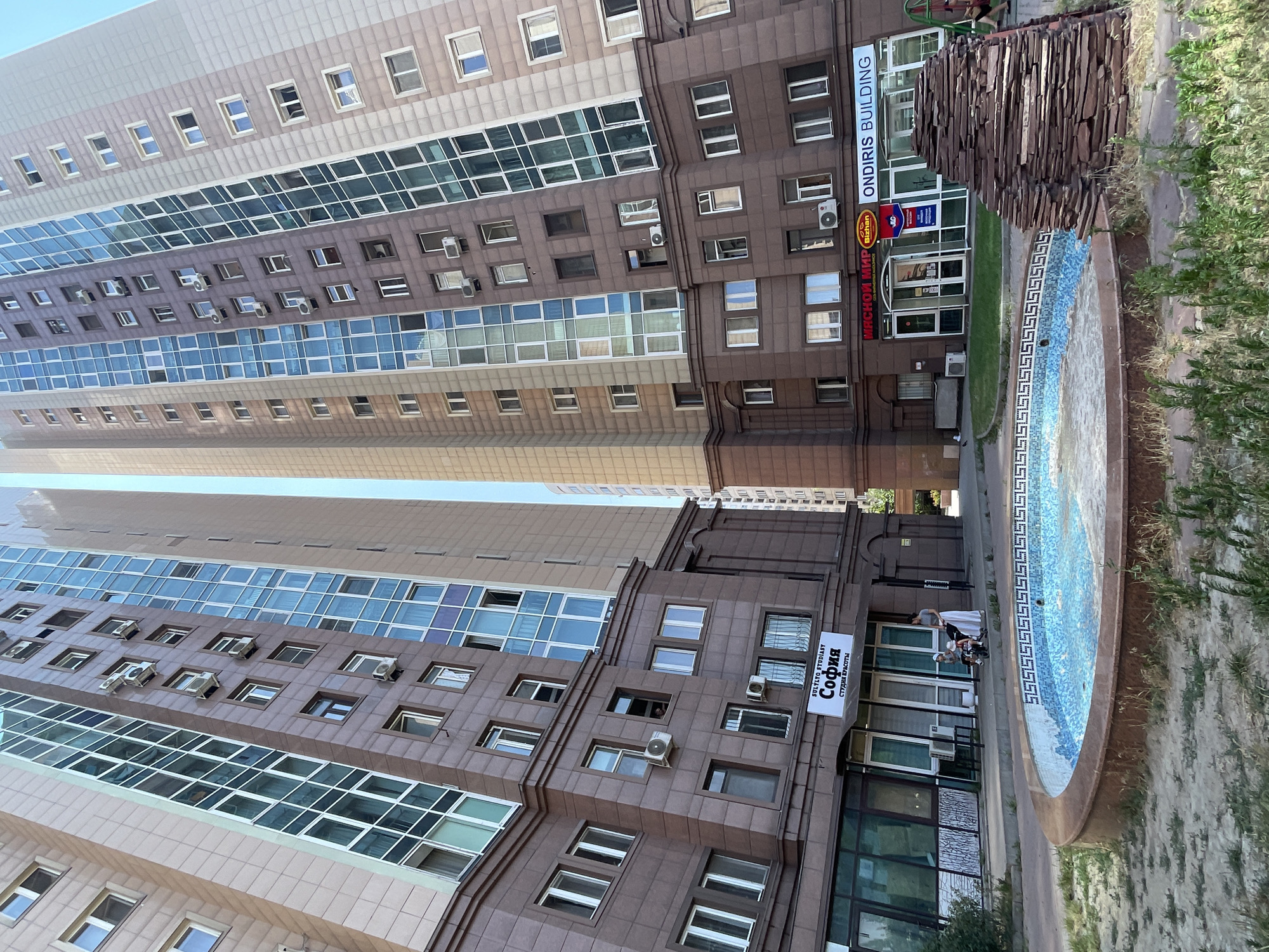 The image shows a street scene in Almaty, Kazakhstan, featuring two tall, modern apartment buildings that dominate the frame. The buildings are clad in brown and beige tiles with numerous windows and air conditioning units visible. In the foreground, a circular fountain with a mosaic tile pattern is partially filled with water. A woman and two children are near the fountain, with the woman pushing a stroller. Several shops are located on the ground floor of one of the buildings, including one with a sign that reads "SOFIA STUDIO". The sky is clear and blue. The area surrounding the fountain is paved with sand and sparse vegetation. A stacked stone sculpture is visible to the right of the fountain.
