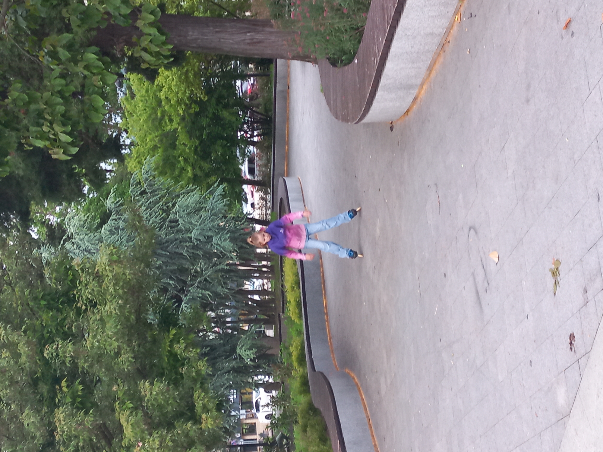 A young person in a purple and pink hooded top and blue jeans is in the air, possibly skateboarding, in what appears to be a paved skate park or public space in Sochi, Russia. The area is bordered by lush greenery including mature trees and shrubs. A concrete structure, perhaps a bench or wall, with a curved edge runs along the right side of the image. The ground is gray and marked with lines suggesting paving stones. In the background, glimpses of buildings and parked cars are visible through the trees.