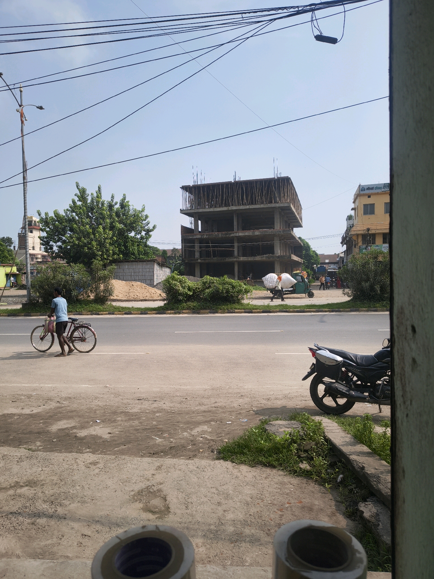 The image depicts a street scene in Purnia, India, with construction and daily life occurring simultaneously. In the foreground, a person is walking a bicycle loaded with pink bags along the side of a paved road. A black motorcycle is parked on the shoulder of the road to the right. In the background, an unfinished multi-story building stands under construction, with scaffolding and exposed concrete. Across the street, an auto-rickshaw is carrying large white sacks, and other people are visible near a yellow building with signage in Hindi. Power lines crisscross the sky, and vegetation, including trees and bushes, frames the scene. The sky is clear and blue, suggesting a daytime setting. The overall impression is one of a bustling urban environment with ongoing development.