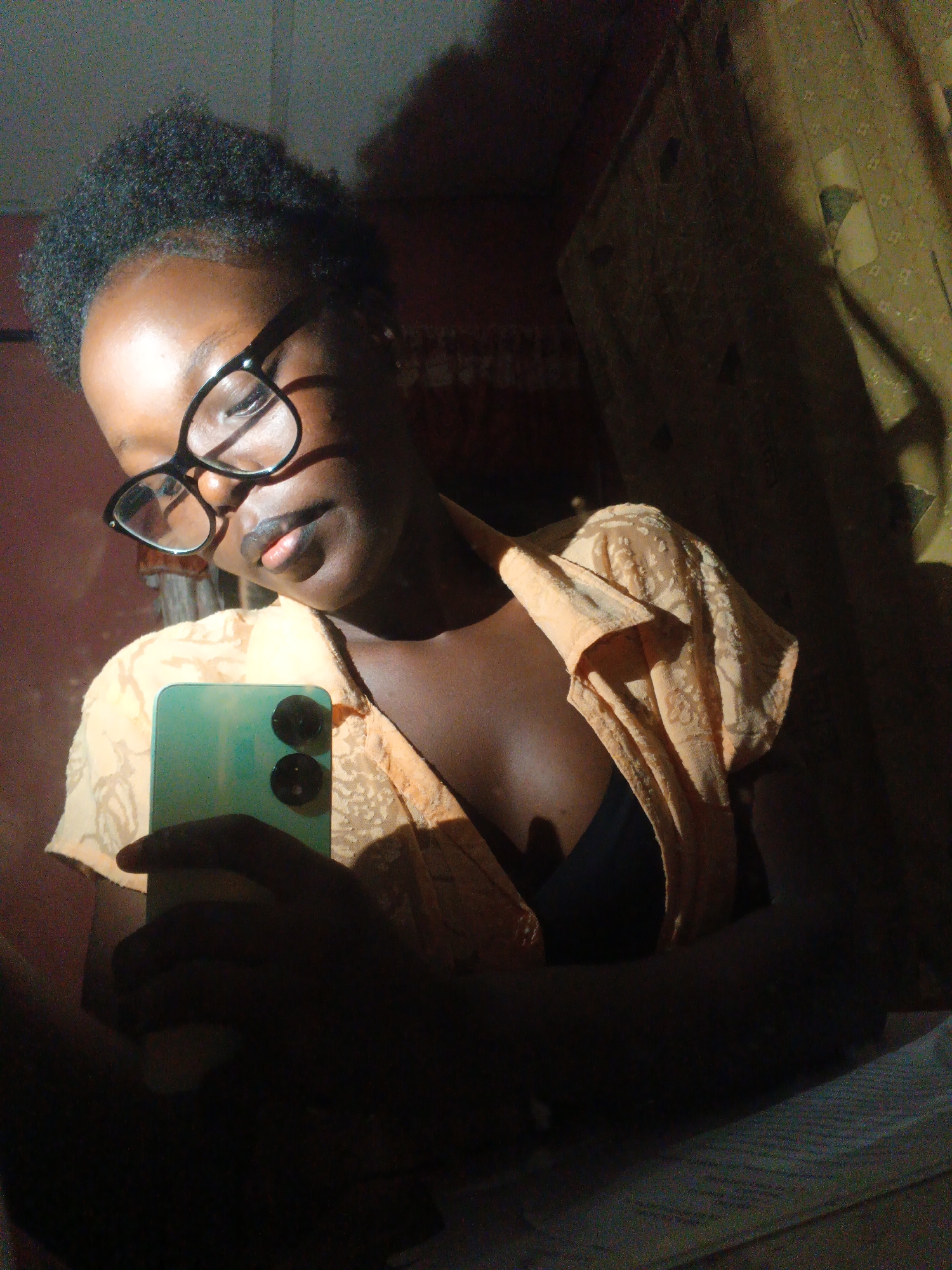 A young Black woman, wearing glasses and a light orange top with a patterned design, is captured in a mirror selfie. She holds a green smartphone in her right hand, which is partially obscured by shadow. The light source appears to be from the right, casting strong shadows across her face and the background. The background consists of a textured wall and what looks like a patterned curtain or tapestry. The image has a grainy quality, typical of low-light smartphone photography. The overall setting suggests an indoor environment, possibly a residential room. The exact location within Onitsha, Nigeria, cannot be determined from the visual information provided.