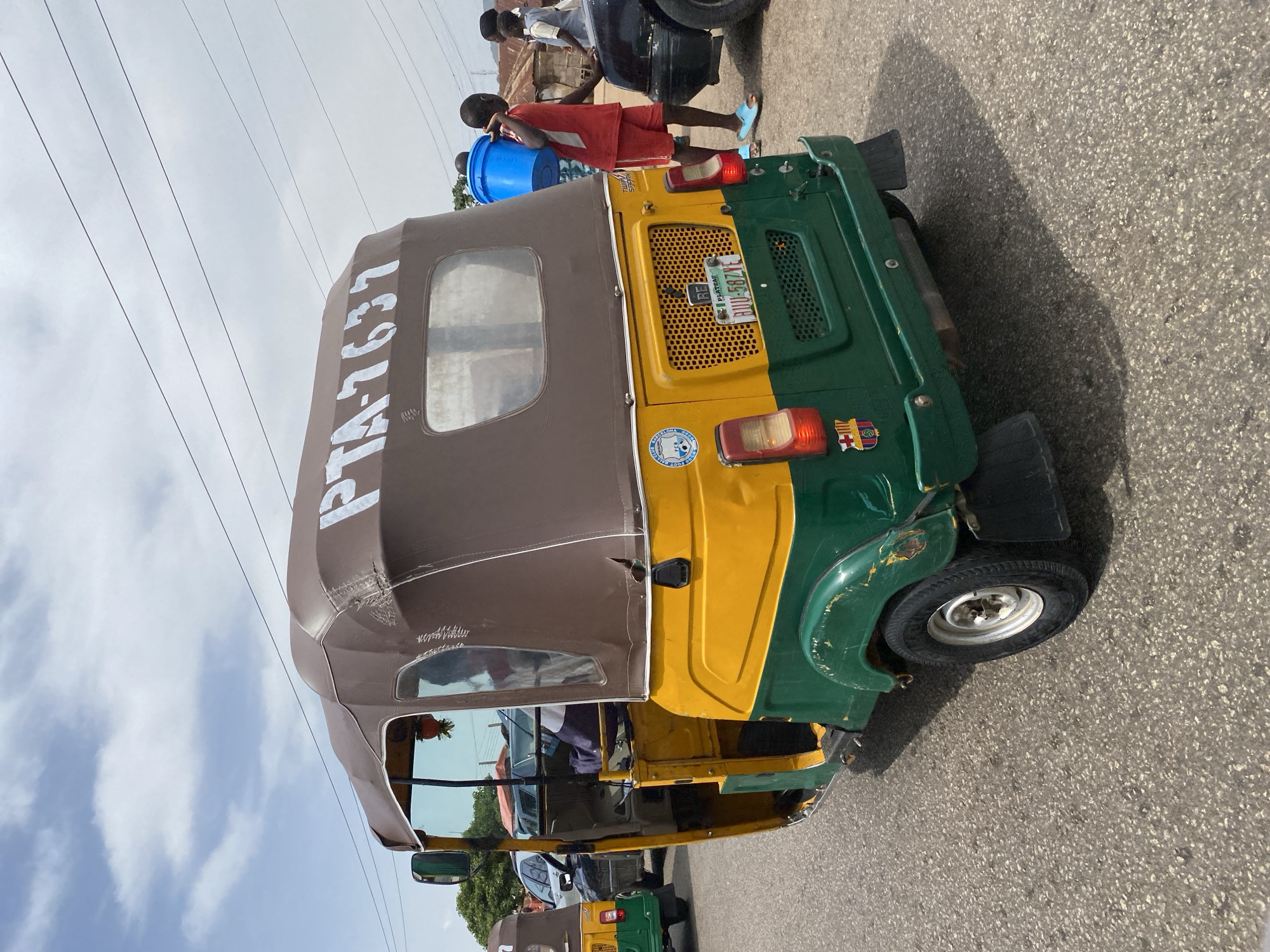 The image shows a three-wheeled vehicle, a Bajaj RE or "Keke Napep," parked on a paved surface. The vehicle is painted in a green, yellow, and brown color scheme. Its license plate reads BU-582YE. The vehicle's roof is labelled PTA-7637. Several people, including a child in red shorts, are standing near the vehicle. A blue plastic container is visible on the rear of the vehicle. The location is identified as Bukuru, Nigeria. The presence of overhead power lines suggests an urban or suburban setting.