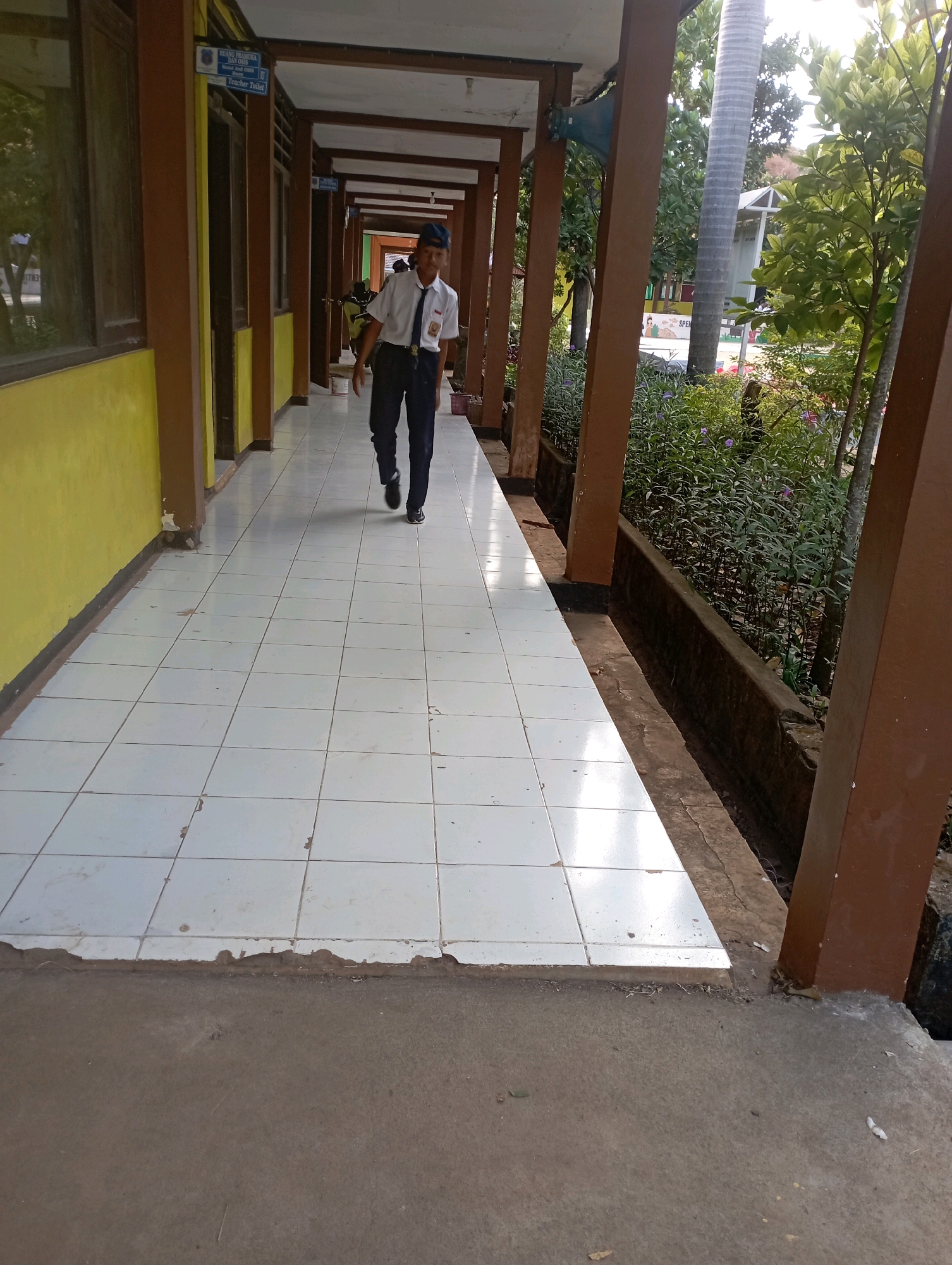 A young male student wearing a white shirt, dark pants, and a blue baseball cap walks down a tiled outdoor hallway. The hallway has brown pillars and is adjacent to a yellow wall on the left and lush greenery on the right. The student appears to be in motion, with his right leg extended forward. The ground is paved with white tiles in a grid pattern, and there is a concrete walkway in the foreground. The scene appears to be in a school or institutional setting, possibly in Banyuurip, Indonesia, given the surrounding vegetation and architectural style. The lighting suggests it is daytime, with sunlight casting shadows.
