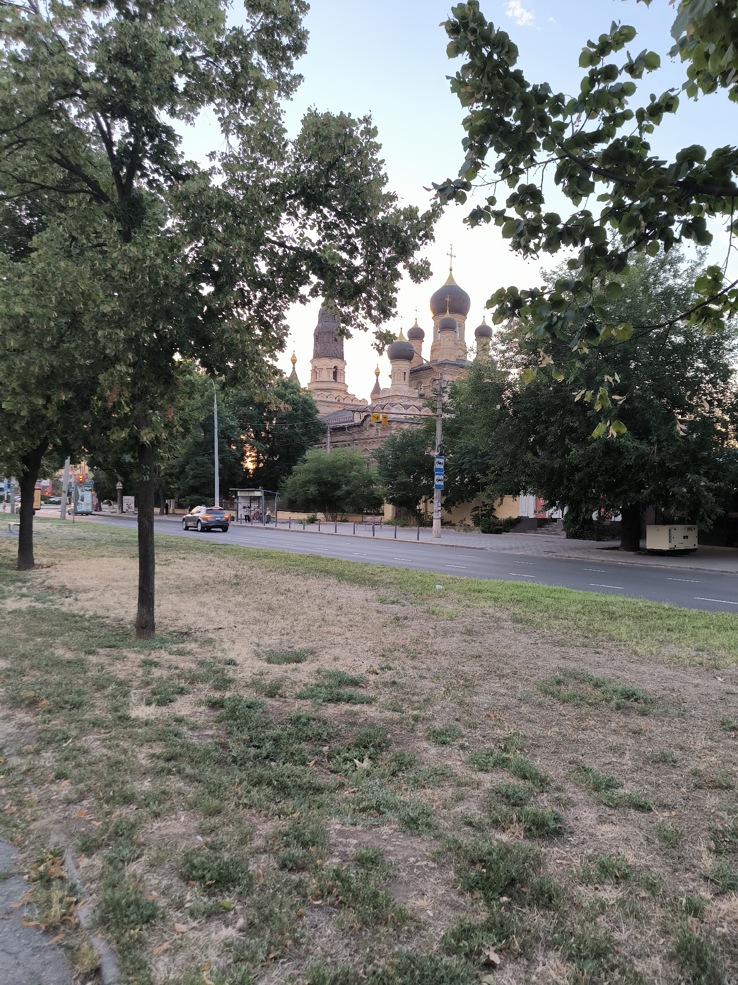 In Mykolaiv, Ukraine, a street scene unfolds with a car driving on the right side of the road. A church with multiple domes, characteristic of Orthodox architecture, stands in the background, partially obscured by trees. The foreground features a grassy median with sparse vegetation and dry patches. A bus stop shelter is visible on the sidewalk. The sky is clear, suggesting daytime.