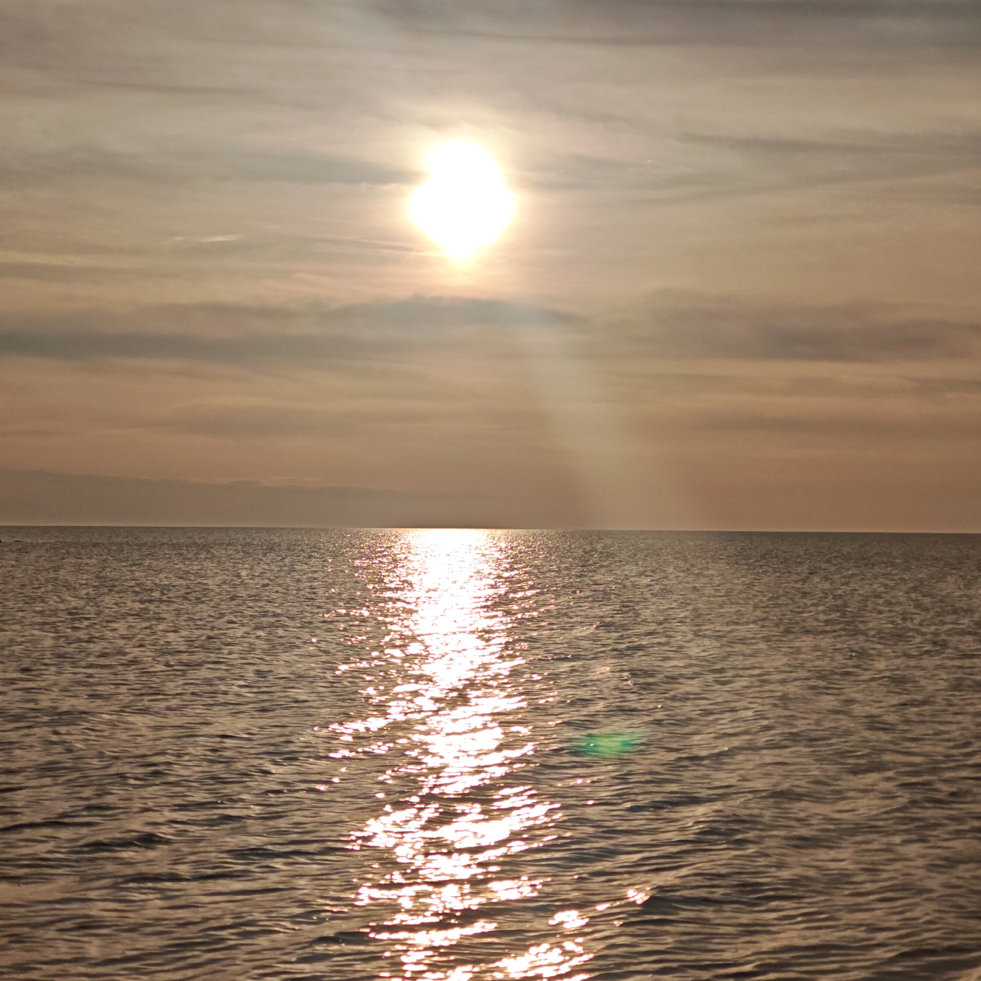 The image shows a serene scene of the sea at sunset in городское поселение Щелкино, Ukraine. The sun, a bright, circular orb, is positioned high in the sky, its light reflecting off the water's surface. The sky is partly cloudy, with layers of grey and beige clouds spread across the horizon. The sea is calm, with gentle waves rippling across its surface. The sun's reflection creates a shimmering pathway of light that extends from the horizon towards the foreground. A slight green lens flare is visible in the lower-right quadrant of the image.