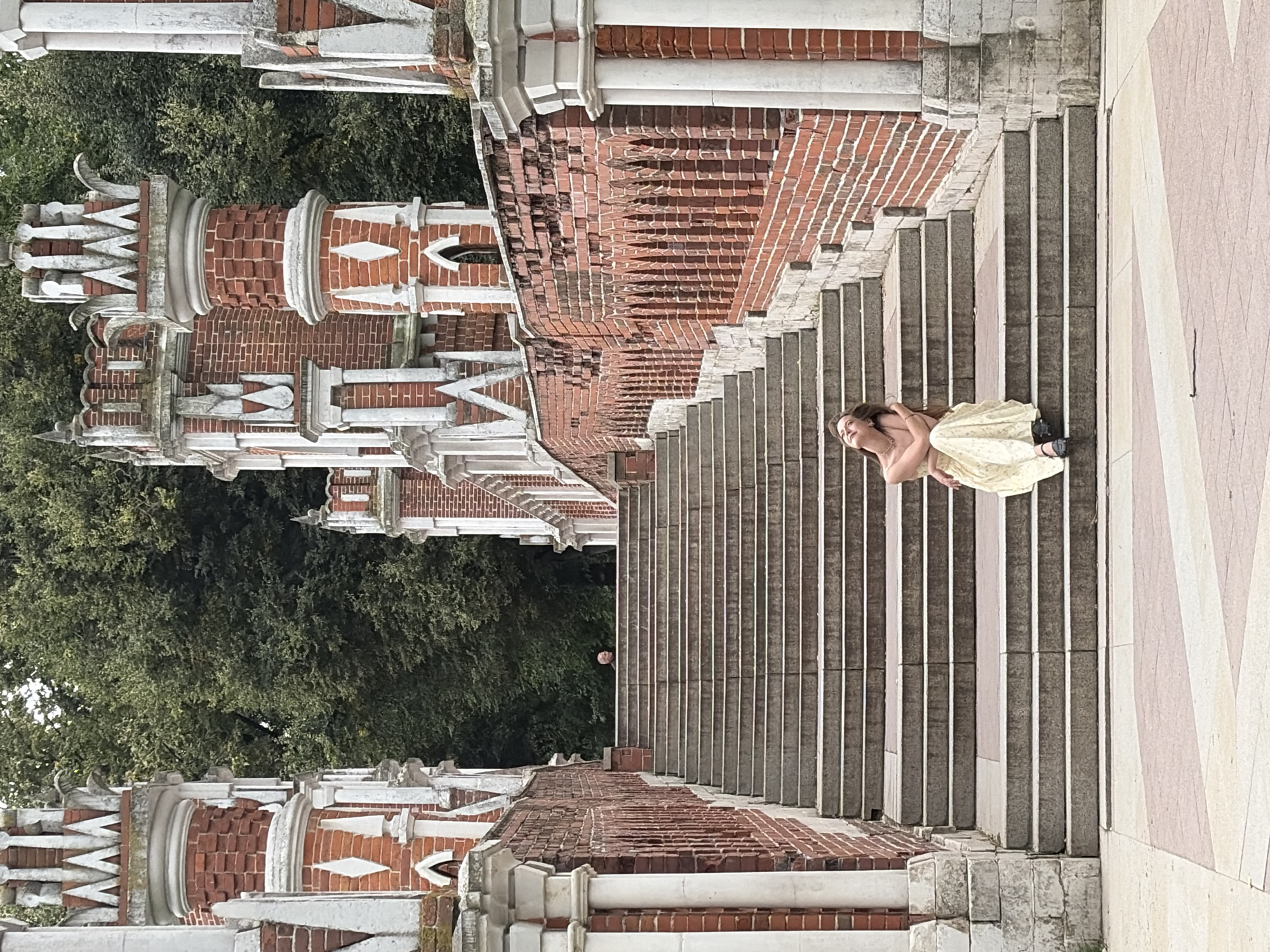 A young woman wearing a light-colored, long, patterned dress and black shoes sits on the steps of a stone staircase. She is leaning back with her head tilted up and eyes closed, her arms around herself. The staircase is flanked by a red brick wall with decorative arches and white stone columns and turrets, characteristic of Russian Neo-Gothic architecture. In the background, lush green trees form a backdrop. The scene appears to be in Tsaritsyno Park in Moscow, Russia, a popular historical and architectural complex.