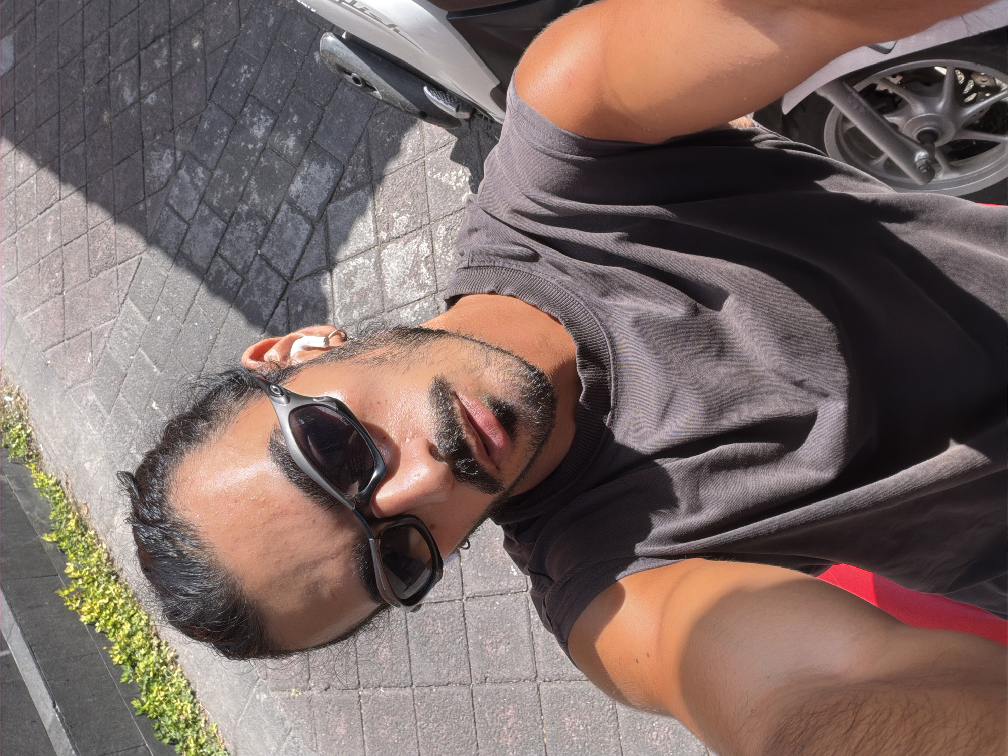 A selfie of a man with dark hair pulled back, wearing dark sunglasses and a gray t-shirt, is taken outdoors on a paved area. The man has a goatee and mustache and is wearing white earbuds and a hoop earring. To the right of the man is a white scooter. The background consists of a grey, patterned stone path, and some green foliage and stone steps to the left. A shadow from an unseen object falls across the paved area. The lighting suggests it is daytime.