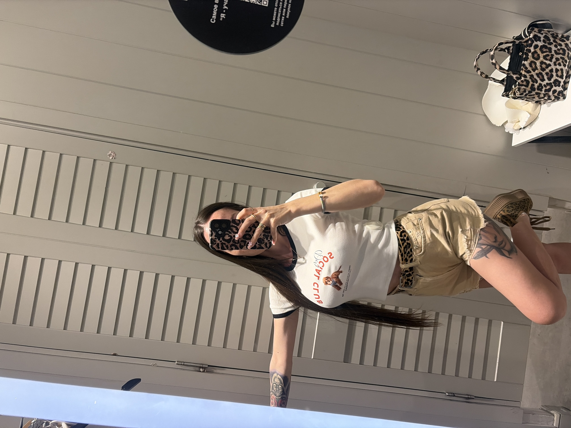 A woman with long dark hair is taking a mirror selfie in what appears to be a fitting room or a section of a retail store, possibly in Saint Petersburg, Russia. She is wearing a white cropped t-shirt with a graphic print of a dog and red text that reads "SOCIAL CLUB", a leopard-print belt, and light beige distressed shorts. She also has tattoos on her left arm and right thigh, a bracelet on her left wrist, and rings on her fingers. A leopard-print handbag and a white item, possibly a hat, are on a shelf to her right. In the background, there is a white wall with horizontal paneling and a circular black sign with white text. The woman is holding a smartphone with a leopard-print case in her right hand.
