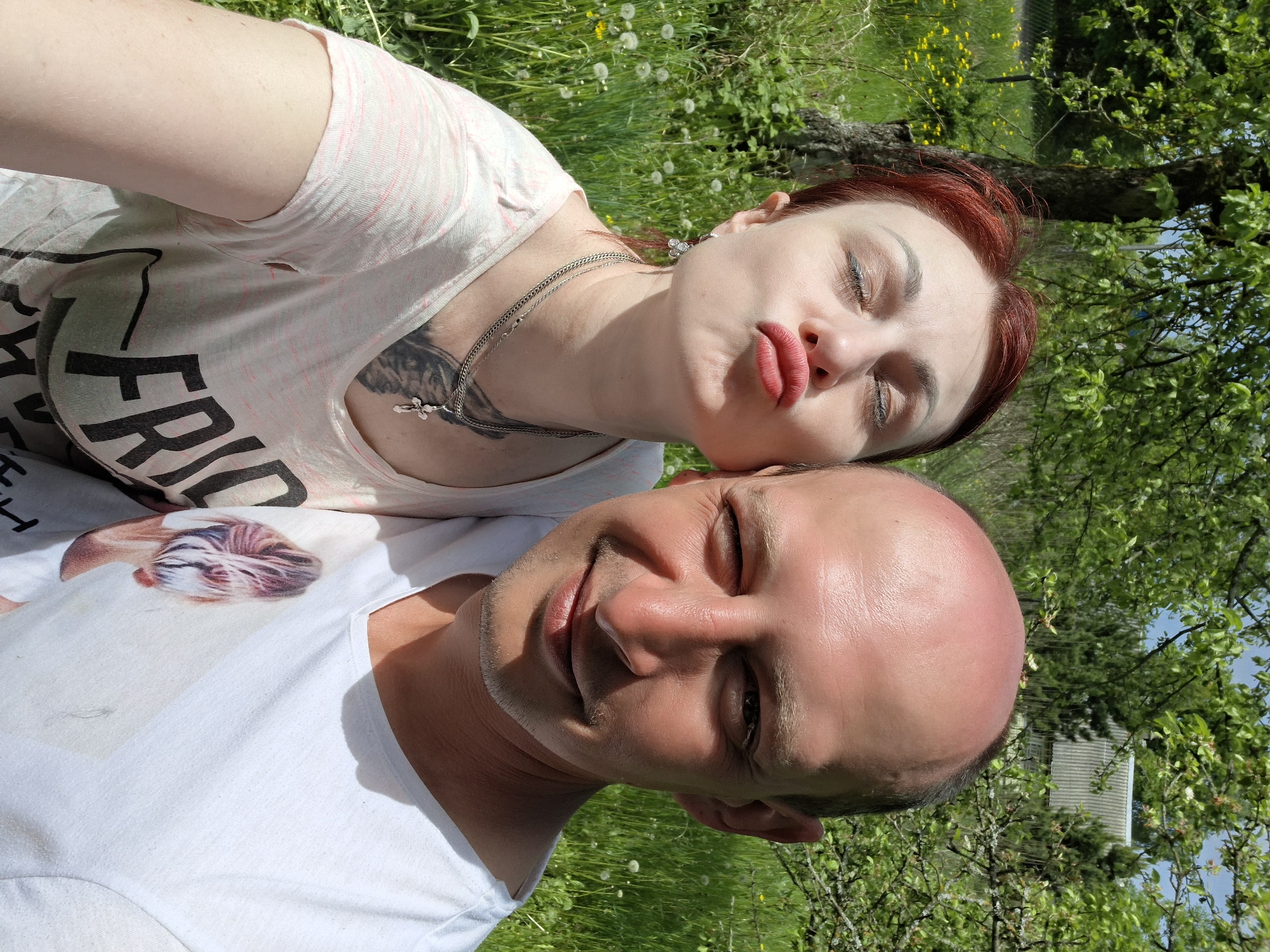 A selfie shows a man and woman in a grassy area with trees. The man is wearing a white t-shirt and is slightly in the foreground, with his head tilted towards the camera. The woman is lying behind him, close to his head. She has red hair and is wearing a light-colored t-shirt with a graphic on it. She is pouting with her eyes closed. The location is Сваткаўскі сельскі Савет, Belarus.
