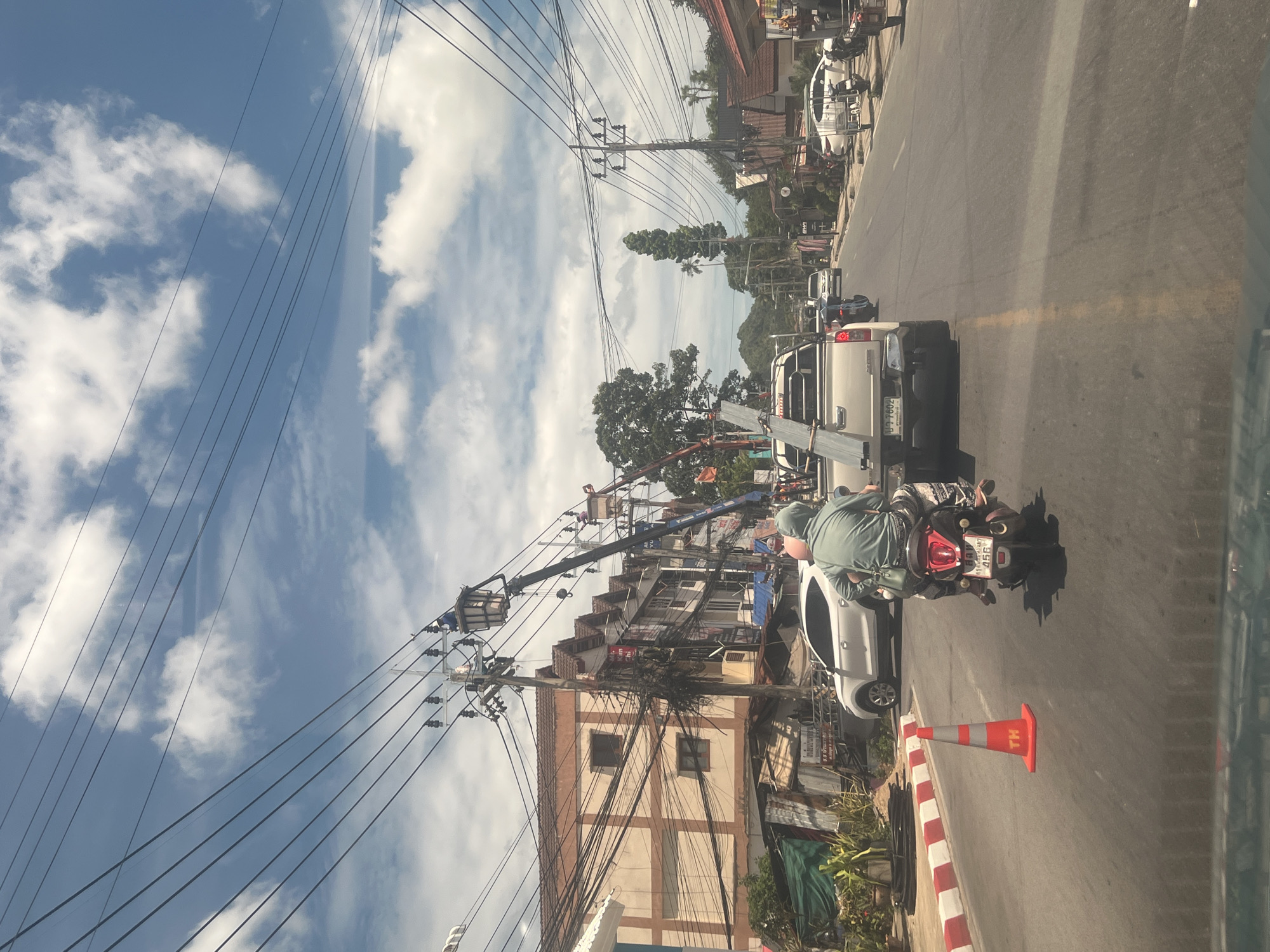 In Ko Samui District, Thailand, on a sunny day with scattered clouds, a street scene unfolds. The road is lined with buildings and utility poles, with a complex network of overhead wires. A white pickup truck is visible, carrying a long metallic object. In front of it, a person on a motorcycle, wearing a light green hooded shirt and patterned pants, is traveling down the street. To the left, a white car is parked. Further down the road, a utility worker in a bucket truck is attending to the power lines. A traffic cone is placed on the side of the road. The buildings in the background appear to be a mix of residential and commercial structures.