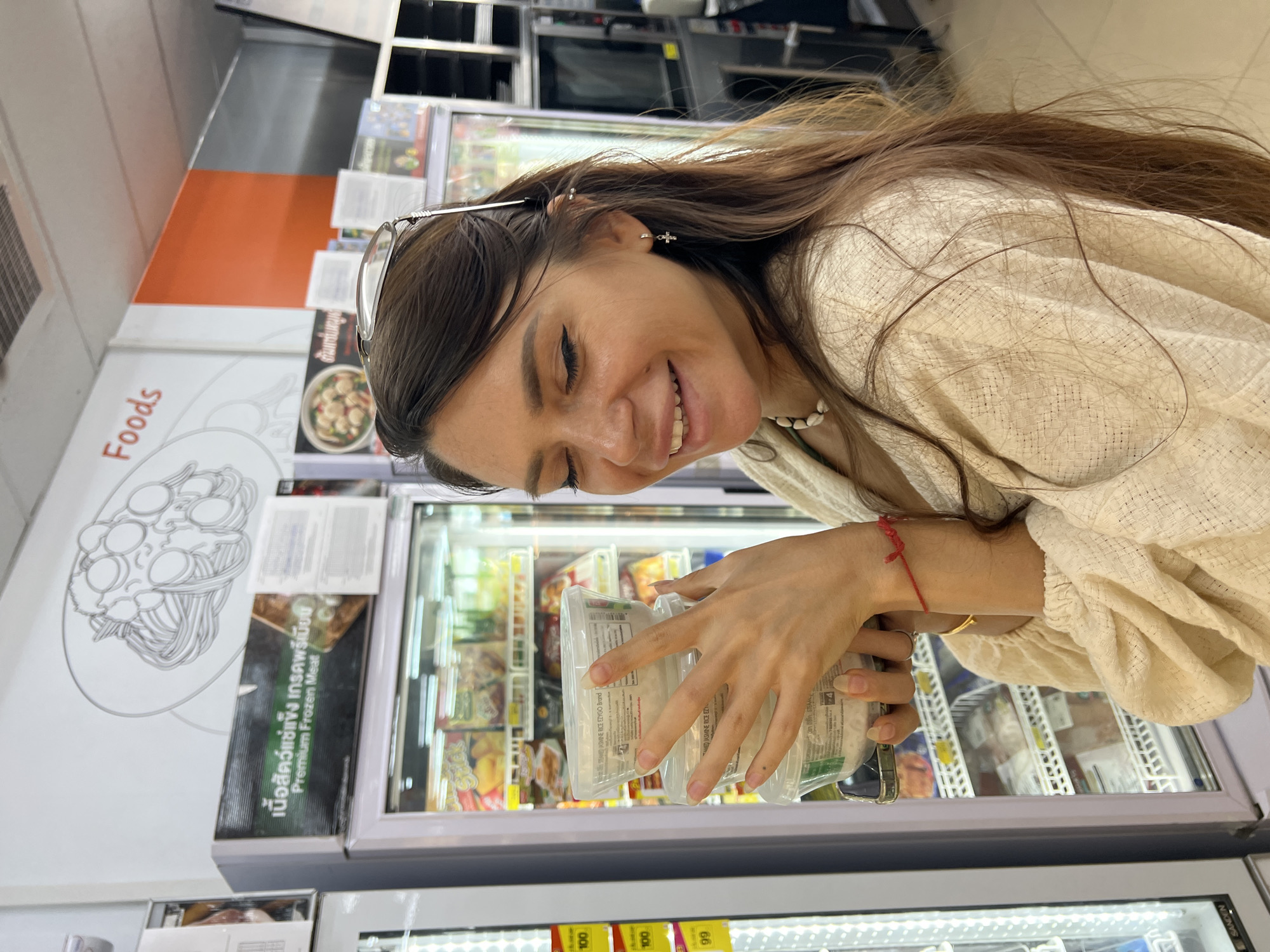 A medium shot captures a woman with long brown hair and closed eyes, smiling while holding three plastic containers of what appears to be rice in a grocery store. She is wearing a cream-colored textured garment and a pearl necklace. A red string is tied around her left wrist, and she has a cross-shaped earring in her left ear. Her eyeglasses are perched on her head. Behind her, a refrigerator case is visible, stocked with various food items. Above the refrigerator, a sign displays text in Thai and English, including "Premium Frozen Meat." The background also shows a wall with a graphic of pasta and the word "Foods" in red. The scene is set in a commercial establishment, likely a convenience store or supermarket, in Ban Bang Rak, Thailand, indicated by the presence of Thai language signage and products.