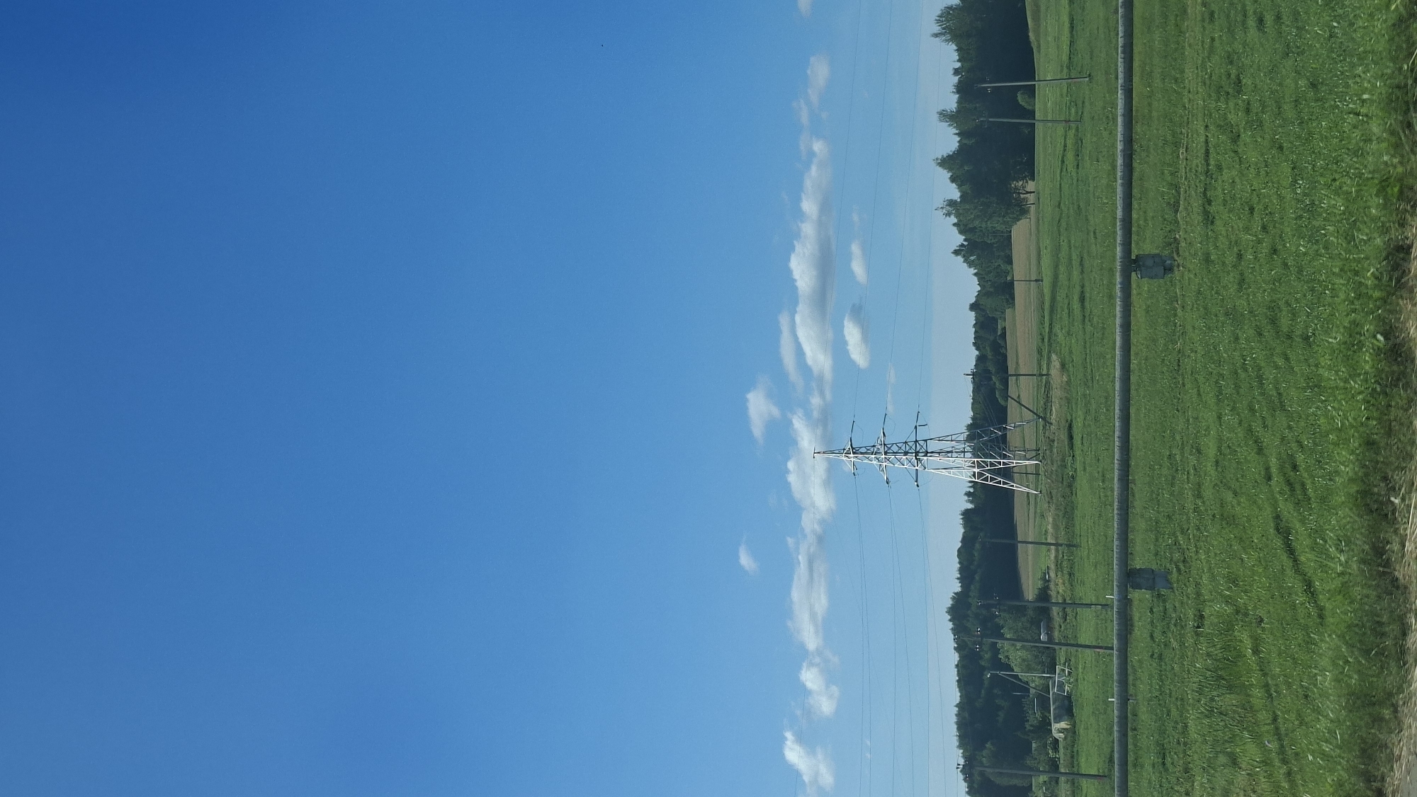 The image depicts a wide-angle view of a rural landscape under a clear blue sky with scattered white clouds. In the mid-ground, a tall, lattice-style electrical pylon stands prominently in a green, grassy field. Power lines extend from the pylon across the frame. A thick, horizontal pipe supported by concrete pillars runs across the foreground. The background features a dense line of trees, and beyond that, rolling hills and fields. The scene appears to be located in or near the city of Myadzyel, Belarus, given the characteristic flat terrain and agricultural landscape often found in the region. There are no people or animals visible in the image.