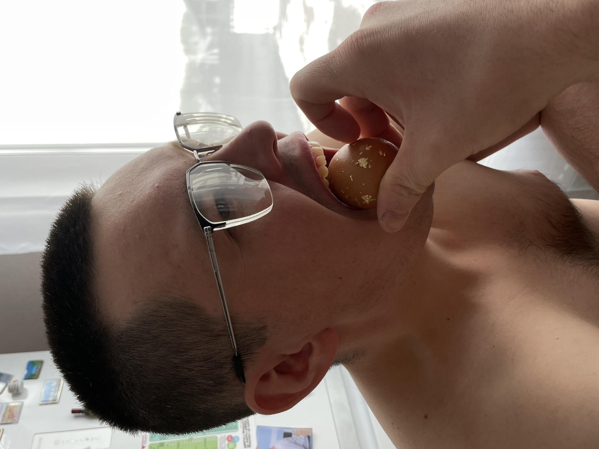 A man with short dark hair and wearing glasses is holding a small, round, brown object with gold leaf flecks near his open mouth. The background is a window with diffused light and a white wall with various small items attached to it. The man is wearing no shirt. The location is not discernible as Подшивалово, Russia.
