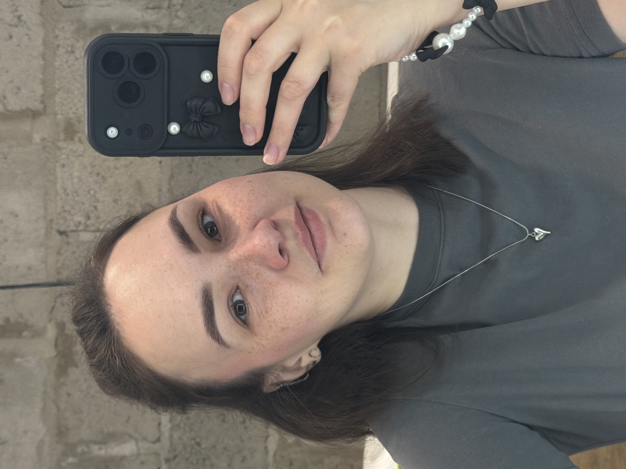 A young, light-skinned woman with brown hair is holding up a black cell phone to take a selfie. The phone case is decorated with a 3D black bow and white pearl-like accents. She is wearing a gray t-shirt and a bracelet with large white beads and small black accents. A silver necklace with a heart pendant is also visible. The background is a blurry, light gray-brown wall. There is no indication that this picture was taken in Chaikovsky, Russia.