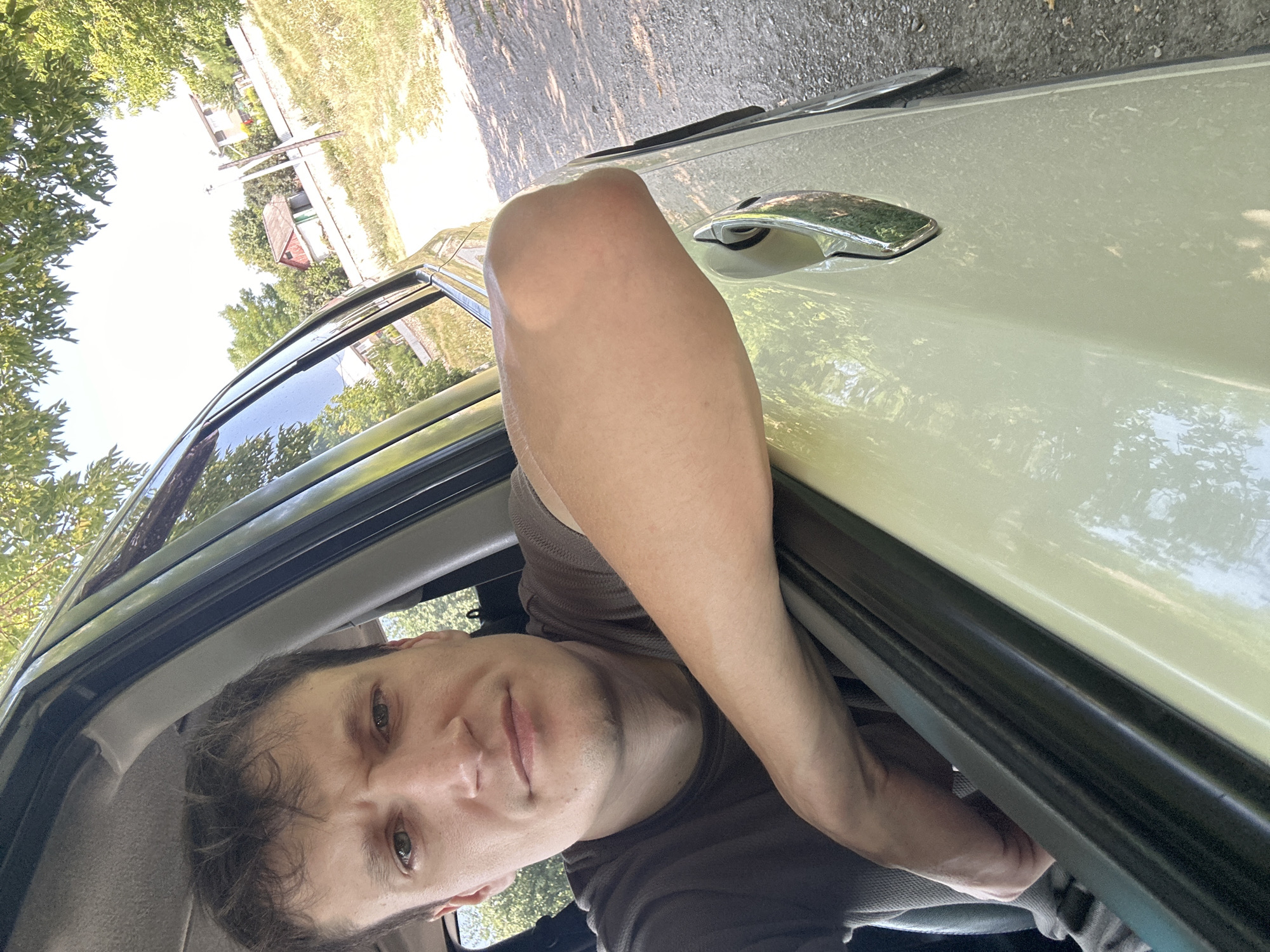 A man is leaning out of a car window with his arm resting on the door. He is looking at the camera with a neutral expression. The car is light-colored and appears to be parked on a dirt road. In the background, there are trees and houses, suggesting a rural or suburban setting. The sun is shining, casting shadows on the ground. The location is identified as Makiv, Ukraine.