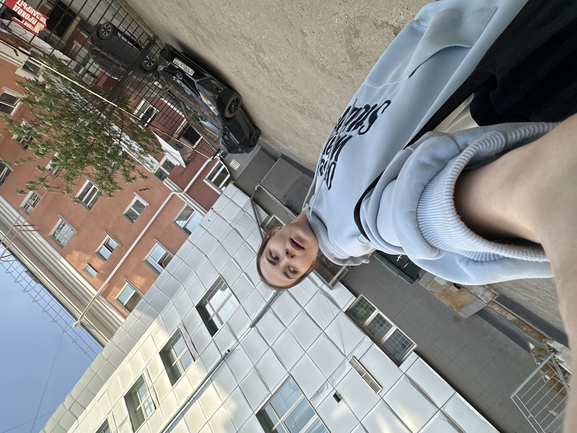The image is a selfie taken in Perm, Russia. The subject is a young woman wearing a light blue hooded sweatshirt with dark lettering and black pants. In the background, buildings are visible, including one with a reddish-brown facade and another with a grid-like white facade. A black car is parked along the street. A small tree is also visible. The image is oriented vertically, with the woman's face appearing towards the bottom of the frame.