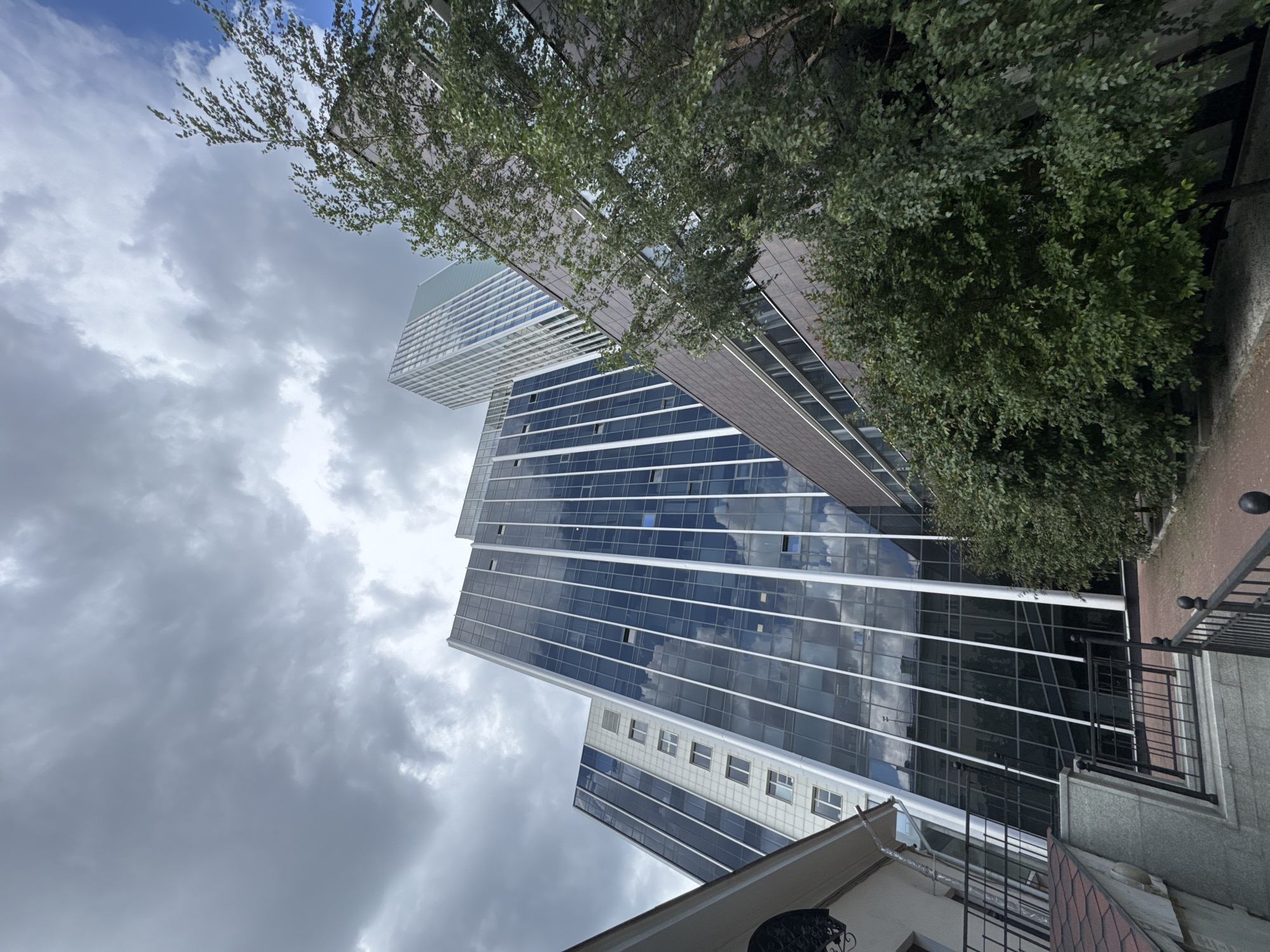 The image depicts a low-angle view of modern buildings and lush green trees under a cloudy sky in Yekaterinburg, Russia. The primary focus is on a tall, angular building with a dark, reflective glass facade, interspersed with vertical white mullions. To its left, a narrower building with a lighter, grid-like facade is visible, featuring rows of rectangular windows. In the background, another taller, sleek skyscraper with a light blue tint can be seen. In the foreground, a dense canopy of green trees partially obscures the lower sections of the buildings. A section of a building with a reddish-brown roof and a white exterior is visible to the left, with a black metal fence in front of it. The overall impression is one of urban development integrated with nature.