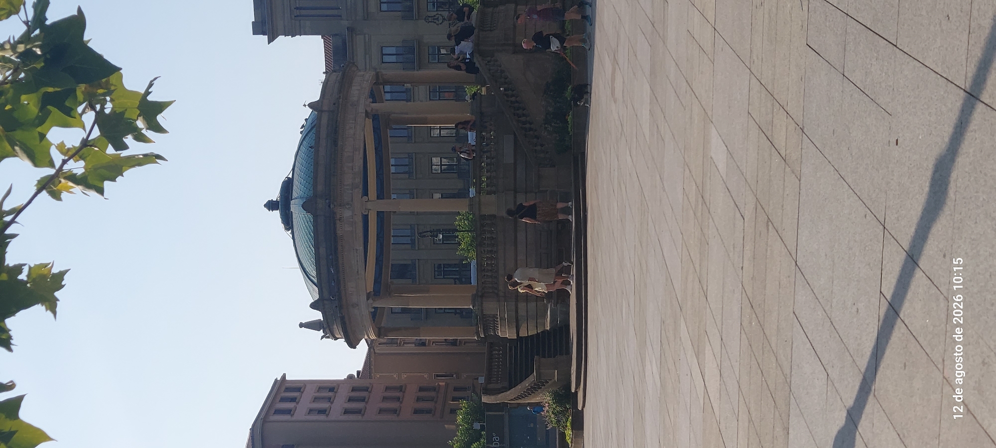 A wide-angle, vertical photograph captures a sun-drenched plaza in Pamplona, Spain, on August 12, 2026, at 10:15 AM. In the center of the frame, a classic bandstand with a domed, green roof stands prominently. The bandstand is supported by a colonnade of weathered stone columns and features intricate stone balustrades and staircases leading up to it. Several individuals are seen scattered around the plaza and on the bandstand's steps. Two people, a man and a woman, walk up the stairs on the left side of the bandstand. Another woman stands near the top of the stairs. On the right side of the bandstand, a woman with dark hair walks a small, dark-colored dog on a leash. Further to the right, a person in a dark shirt and shorts walks away from the camera, carrying a stick. In the background, a multi-story building with numerous windows is visible behind the bandstand. To the left, a tall, pinkish-brown building with many windows lines the edge of the plaza. The ground of the plaza is paved with large, square stone tiles, and a long shadow from an unseen object stretches across it from the left side of the frame. The sky is clear and blue, with a few green leaves from a tree visible in the upper portion of the image.