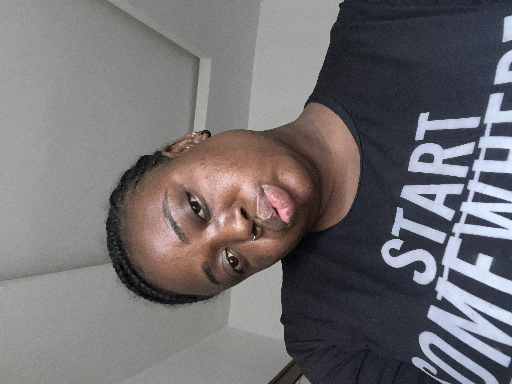 An eye-level close-up shot features a person with dark skin, likely female, looking directly at the camera with a slight pout. Their head is tilted to the left, and they are wearing a black top with white text. The text on the shirt reads "START SOMEWHERE," with parts of the words "START" and "SOMEWHERE" visible. The person has braided hair, specifically cornrows, visible on the side of their head, and a silver ring piercing in their left nostril, along with a small gold-colored earring in their left ear. The background is a plain, light-colored wall, suggesting an indoor setting. The lighting appears natural and bright, highlighting areas of the person's skin. The overall image captures a casual selfie moment. The location context for this image is Jos, Nigeria.