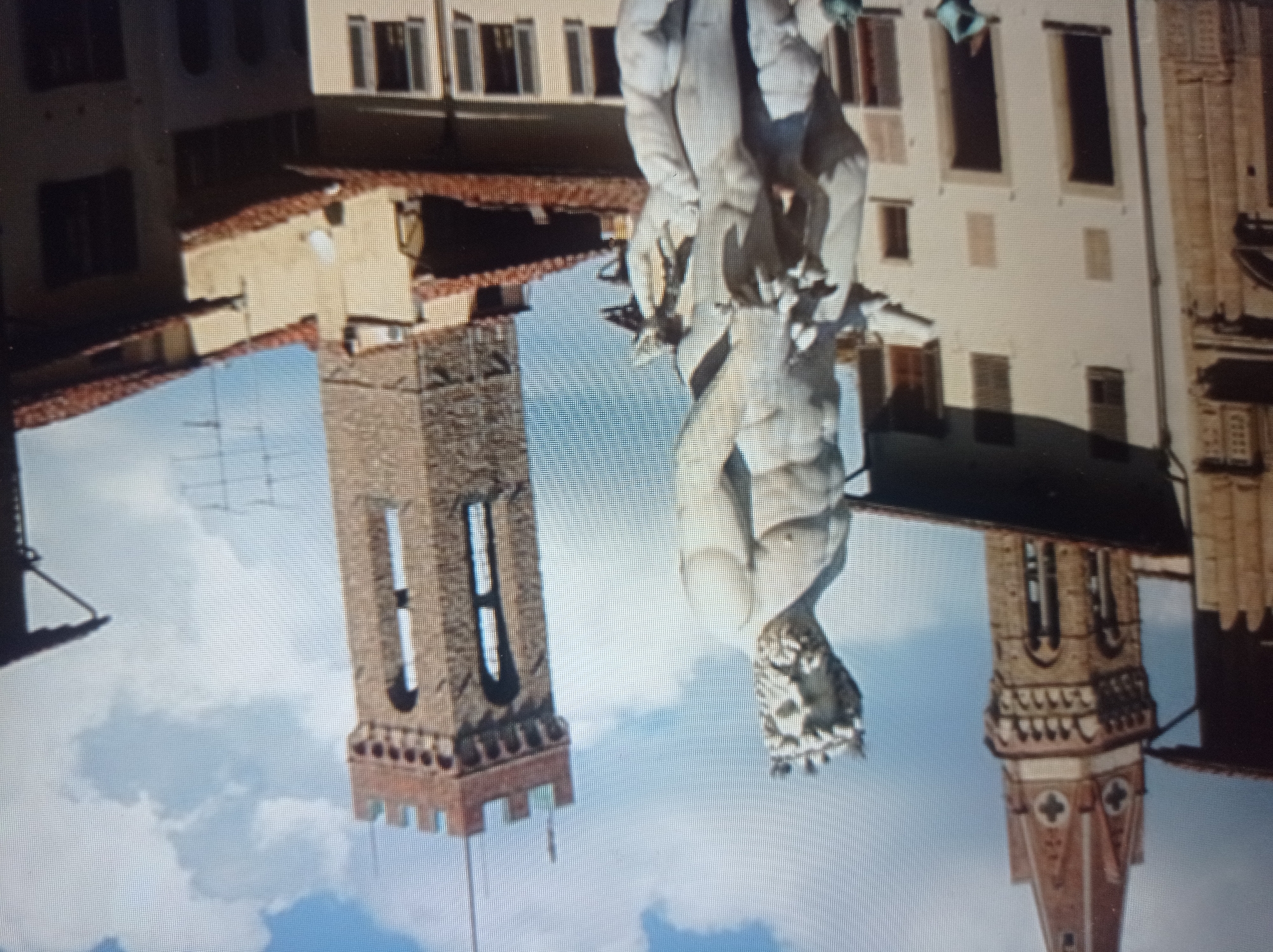The image is a medium shot of the Fontana dei Navigli in Florence, Italy. The statue depicts Neptune, the Roman god of the sea, surrounded by other figures. The statue is made of white marble and is located in a public square. In the background, there are several buildings, including a tall clock tower and other historical structures. The sky is blue with scattered clouds. There are no people in the immediate foreground of the image. The scene is well-lit, suggesting it is daytime.