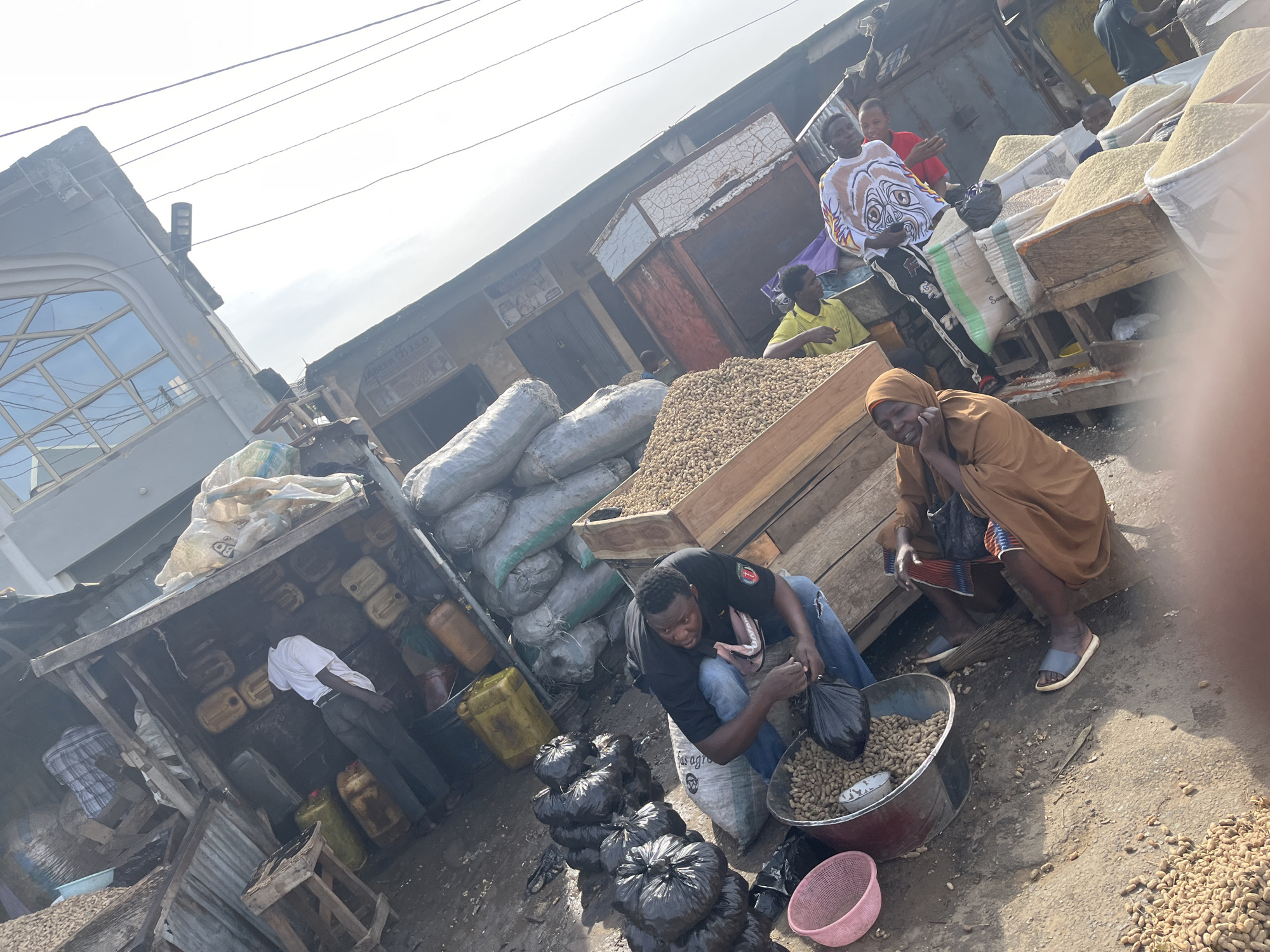 The image depicts a bustling market scene, likely in Jos, Nigeria, with several individuals engaged in trade. In the foreground, a man in a black t-shirt and blue jeans is crouched beside a large metal basin filled with peanuts. He appears to be sorting or bagging the peanuts, with a black plastic bag in his hand. Nearby, a woman in a brown headscarf and traditional attire sits on a wooden bench, looking towards the left. To her right, a broom leans against the bench. Behind them, several large sacks are piled up, some appearing to contain grains.
In the mid-ground, a man in a yellow shirt is visible, and further back, a man in a white t-shirt with an elaborate graphic design and another man in a red t-shirt are present. The environment suggests an outdoor market with makeshift stalls constructed from wood and corrugated metal. Various containers, including yellow jerrycans, are stacked in the background, and large grey sacks are also visible. The ground is dirt and appears somewhat wet in places. The overall impression is one of active commerce in a vibrant market setting.