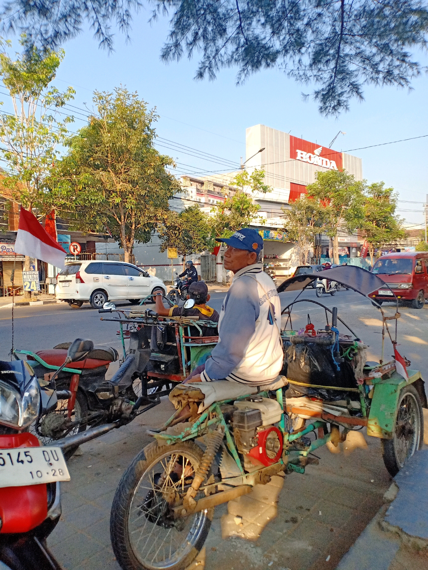 An outdoor, eye-level, medium shot shows a man sitting on a three-wheeled vehicle. The man is wearing a blue baseball cap and a light blue and white jacket with "4" on the back. He is looking to his left. The three-wheeled vehicle is green and has a canopy. It is parked on a sidewalk next to a street. In the background, there is a white car, a red van, a motorcycle, and buildings, including one with a large "HONDA" sign. Trees and power lines are also visible. The Indonesian flag is attached to a pole on the left side of the image. The sun is shining, casting shadows on the ground.