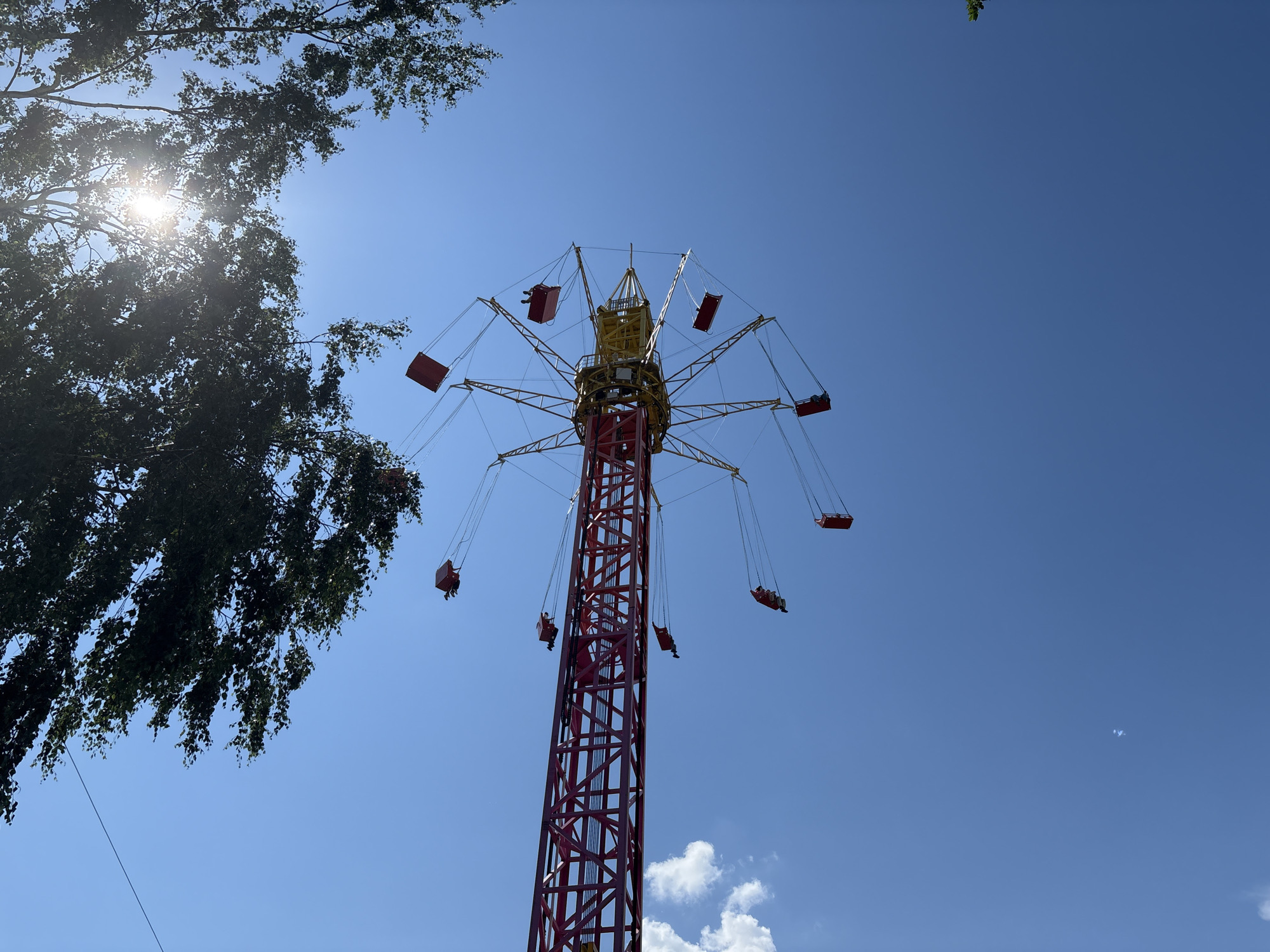 The image shows a tall, red amusement park ride with yellow accents against a clear blue sky. The ride features several red, box-like seats suspended from cables, with people visible in some of the seats. The ride appears to be rotating, as indicated by the position of the seats. In the upper left corner, there are dark green tree branches with sunlight filtering through them. The scene is an outdoor amusement park. The location is in the city of Izhevsk, Russia.
