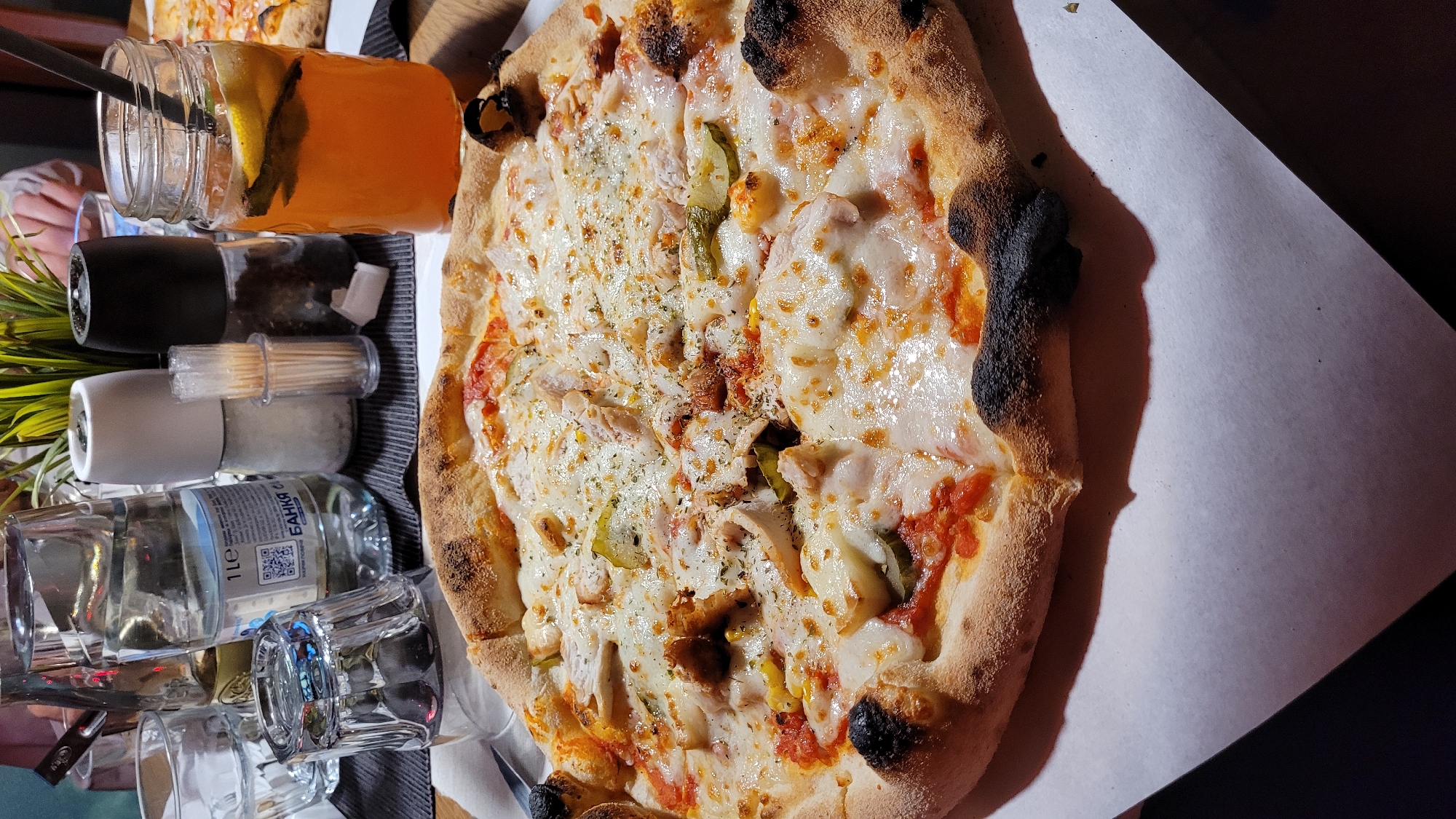 This image presents a top-down view of a table set for a meal, likely in a restaurant or cafe in Varna, Bulgaria. The main subject is a large, irregularly shaped pizza with a thick, partially charred crust, indicating a high-temperature bake. The pizza is generously covered with melted white cheese, a base of red tomato sauce, pieces of cooked chicken, and slices of pickled cucumbers. It is also sprinkled with dried herbs, possibly oregano or thyme, and a few kernels of corn are visible. The pizza rests on white paper or cardboard, placed on a dark surface, possibly a serving board or a striped placemat.
To the left of the pizza, several items are arranged. A clear glass jar, resembling a mason jar, contains an orange beverage, likely iced tea or juice, garnished with slices of lemon or lime and a dark straw. Below this, two shakers, one black and one white, are visible, presumably for salt and pepper. A clear plastic container filled with wooden toothpicks sits nearby. Further to the left, a clear glass bottle with a white label is seen, bearing the Cyrillic text "БАНКА С" and "1Le", along with a QR code, consistent with Bulgarian language use. Several empty clear drinking glasses or tumblers are also present, with one in the foreground showing part of a fork or knife handle. A small artificial green plant adds a decorative element.
The scene depicts a casual dining activity. The lighting is warm and appears to be artificial, suggesting an indoor setting, possibly during the evening. Partially visible hands in the background and lower left corner indicate the presence of diners, although no faces are shown. The image conveys a sense of a relaxed and enjoyable meal.