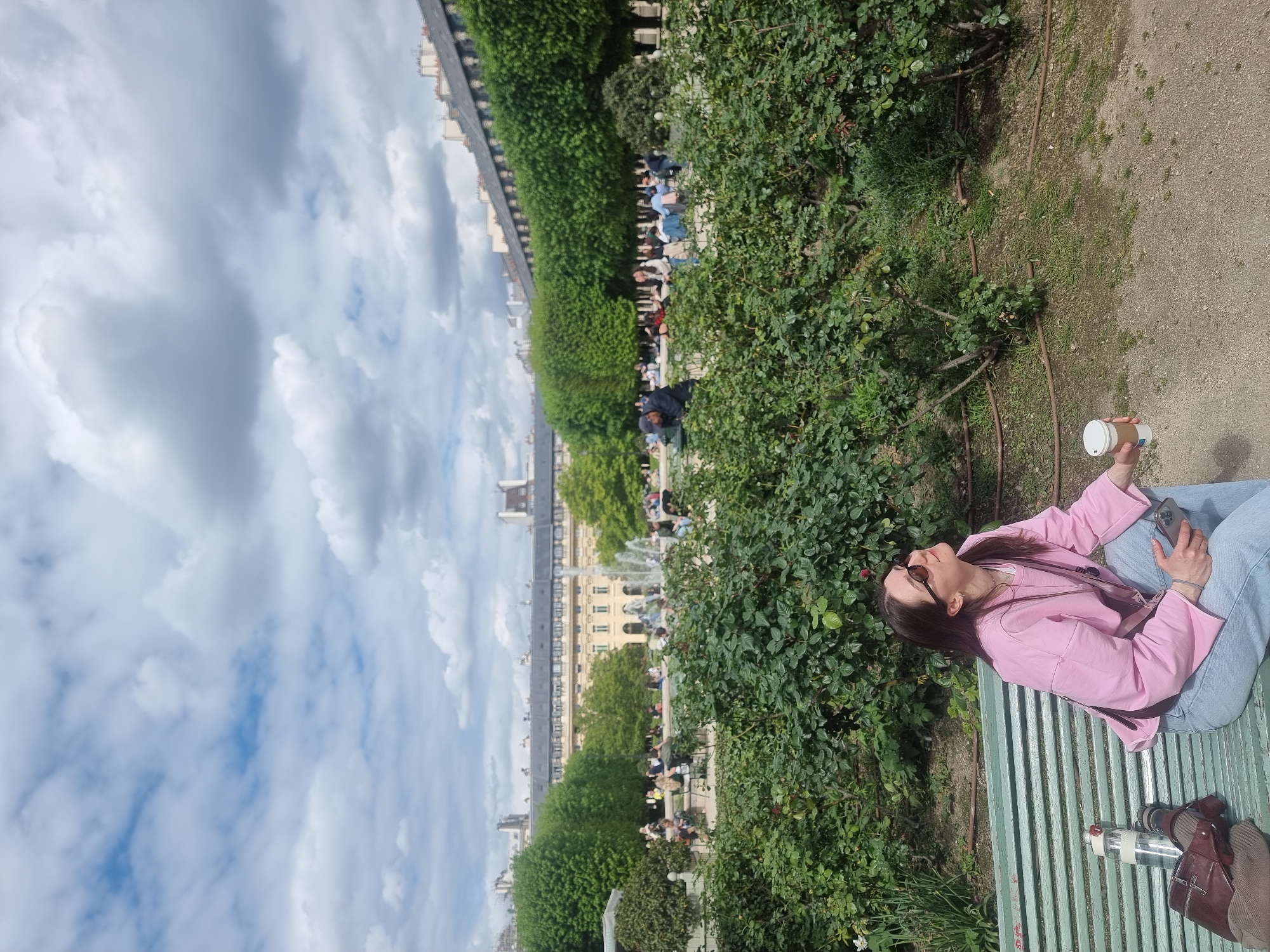 A young woman is shown relaxing on a green slatted park bench in what appears to be a formal garden or park. She is wearing a pink jacket, light blue jeans, and sunglasses perched on her head, with long dark hair cascading down. Her head is tilted upwards, eyes closed, seemingly enjoying the bright daylight. In her right hand, she holds a white paper coffee cup, and her left hand rests on her lap, holding a smartphone. Next to her on the bench, there is a brown leather bag and a clear water bottle.
The setting features lush green bushes and ground cover in the immediate foreground and midground, separating the bench from a dirt or gravel path. In the background, beyond more greenery, a large fountain sprays water, surrounded by other people sitting and strolling. Further in the distance, a long, classical European building with numerous windows and a dark roofline extends across the horizon, characteristic of Parisian architecture (e.g., Tuileries Palace flanking the Tuileries Garden). The sky above is mostly cloudy with large white and grey clouds, though patches of blue are visible. The overall scene depicts a leisurely moment in a public park on a cloudy but bright day. No specific text is clearly legible on any objects.