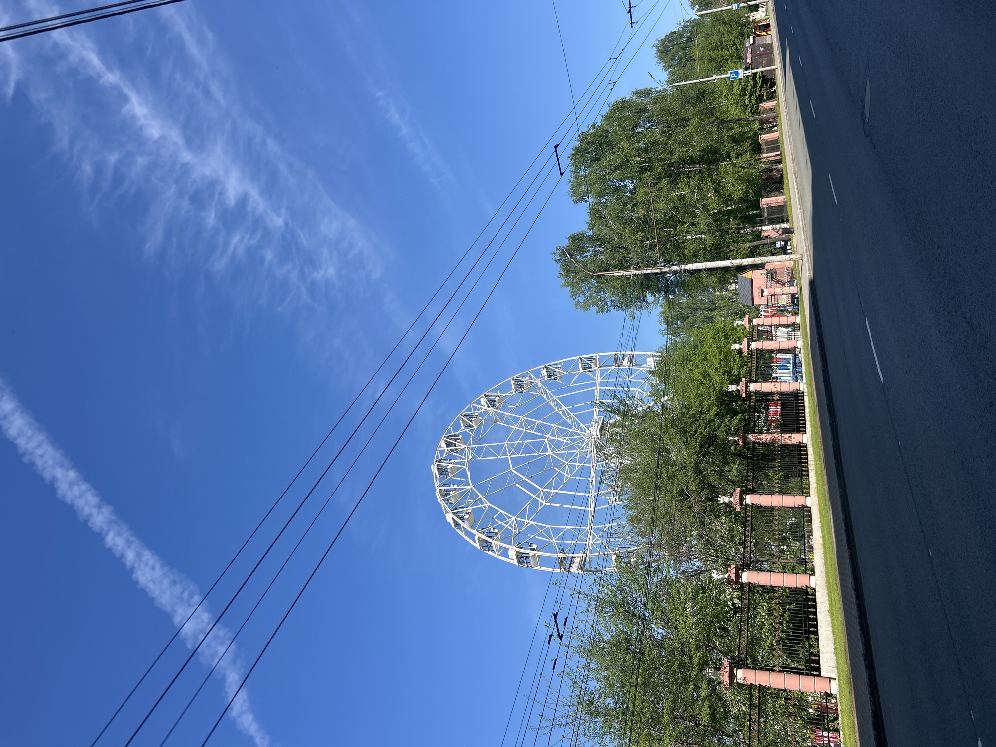 The image shows a Ferris wheel in Izhevsk, Russia, viewed from street level. The wheel is partially obscured by trees. Power lines cross the clear blue sky above. A road runs along the right side of the frame, bordered by a decorative fence.