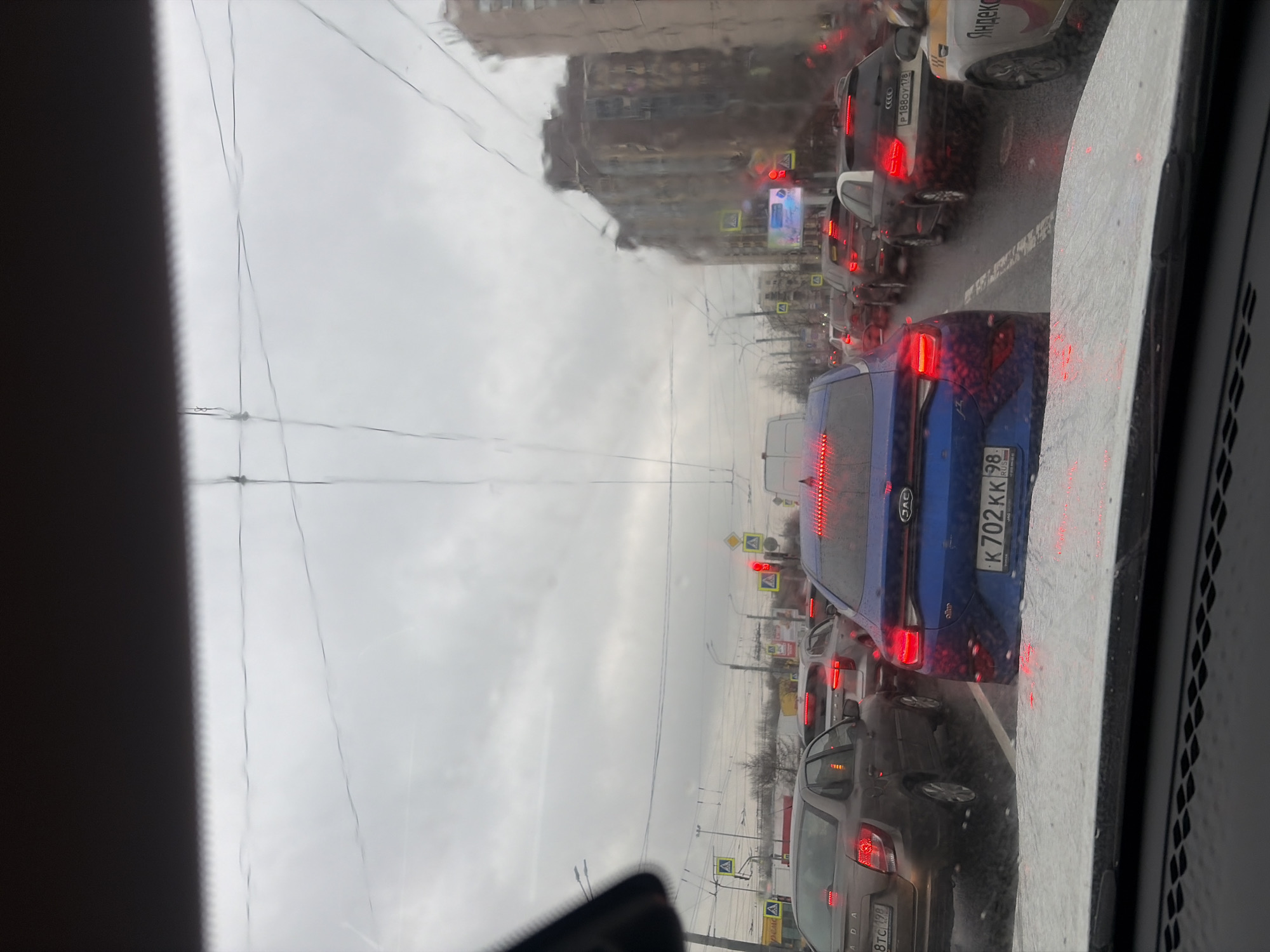 The image shows a view from inside a car, looking out at a traffic jam in Saint Petersburg, Russia. The weather is overcast. A blue car is directly in front, with its license plate partially visible, reading "K702KK 98". Other cars, including a Yandex Taxi, are also visible in the congestion. A building is visible in the background behind the cars, suggesting a dense urban environment. Traffic lights are visible above the cars.