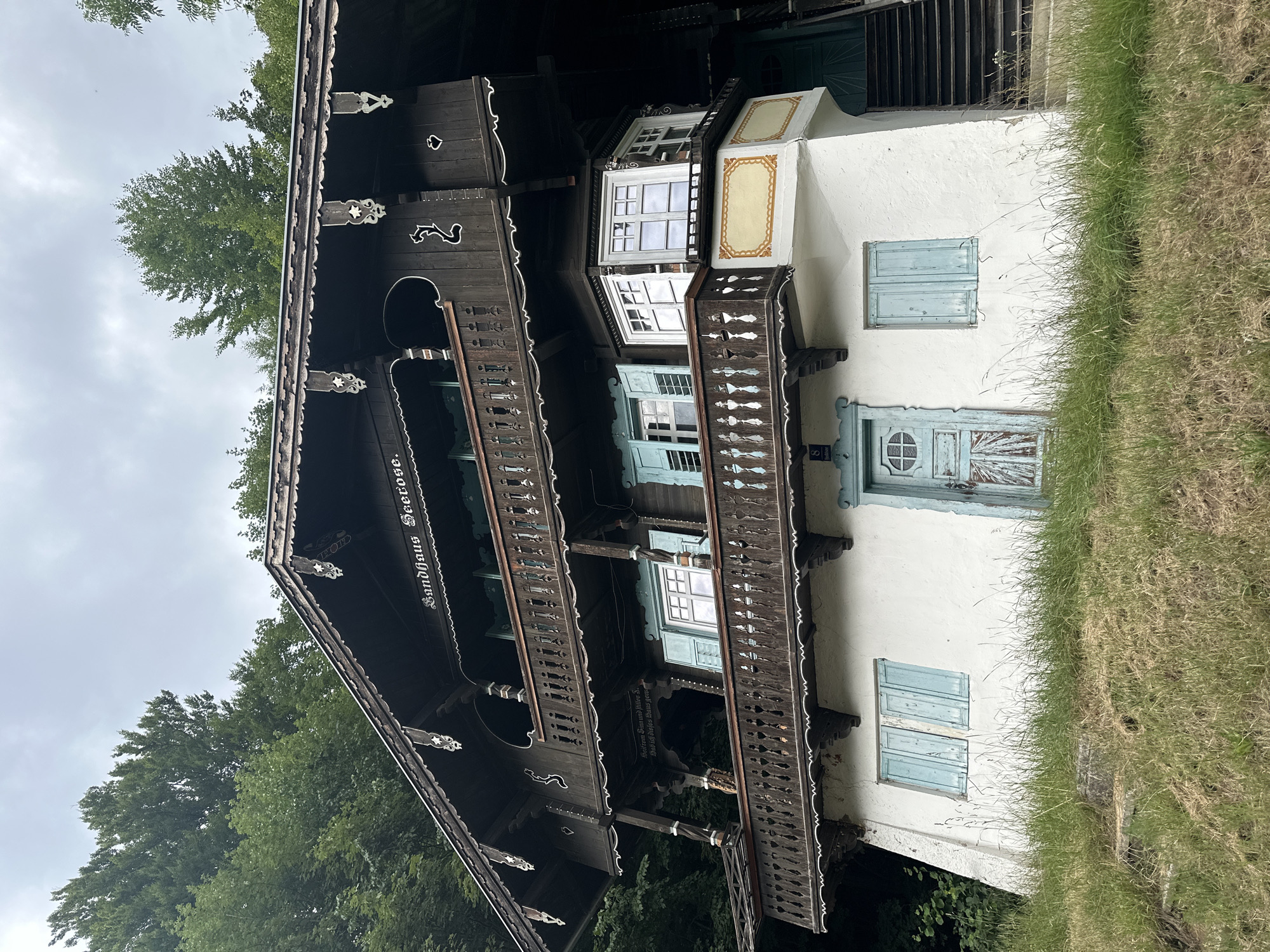 The image shows a view of the "Seerose" guesthouse in Kufstein, Austria, a picturesque town nestled in the Tyrolean Alps. The guesthouse is a traditional Austrian wooden house, characterized by its dark wood facade adorned with intricate carvings and white decorative trim. It features multiple balconies with wooden railings, and light blue shutters on some of the windows. The lower portion of the house is plastered in white, with a light blue door and a window with similar blue shutters. The guesthouse is surrounded by lush green trees, suggesting a natural, possibly mountainous, setting. The sky is overcast, contributing to a somewhat subdued lighting. The foreground is covered in green grass, with a few stone steps leading up to the house, indicating a slightly inclined terrain. There are no people visible in the image.