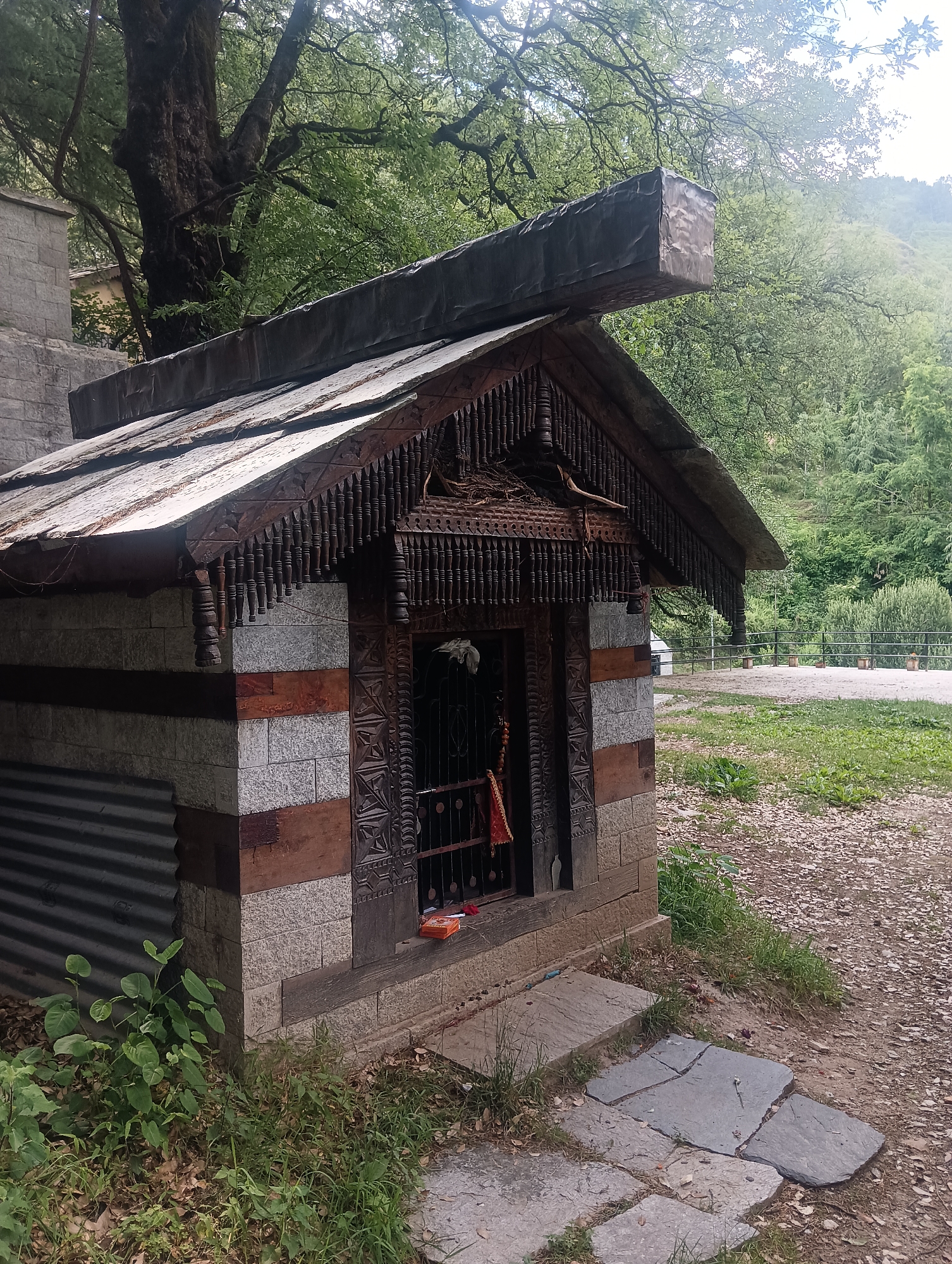The image depicts a small, rustic temple constructed of stone and wood, located in Majhach, India. The temple features an ornate wooden facade with intricate carvings and a tin roof. A large tree with dense foliage stands prominently behind the temple. The surrounding environment is lush and green, with a gravel pathway and some stone slabs leading to the temple entrance. The overall scene suggests a tranquil, natural setting, likely in a rural or mountainous area. There are no people visible in the image.