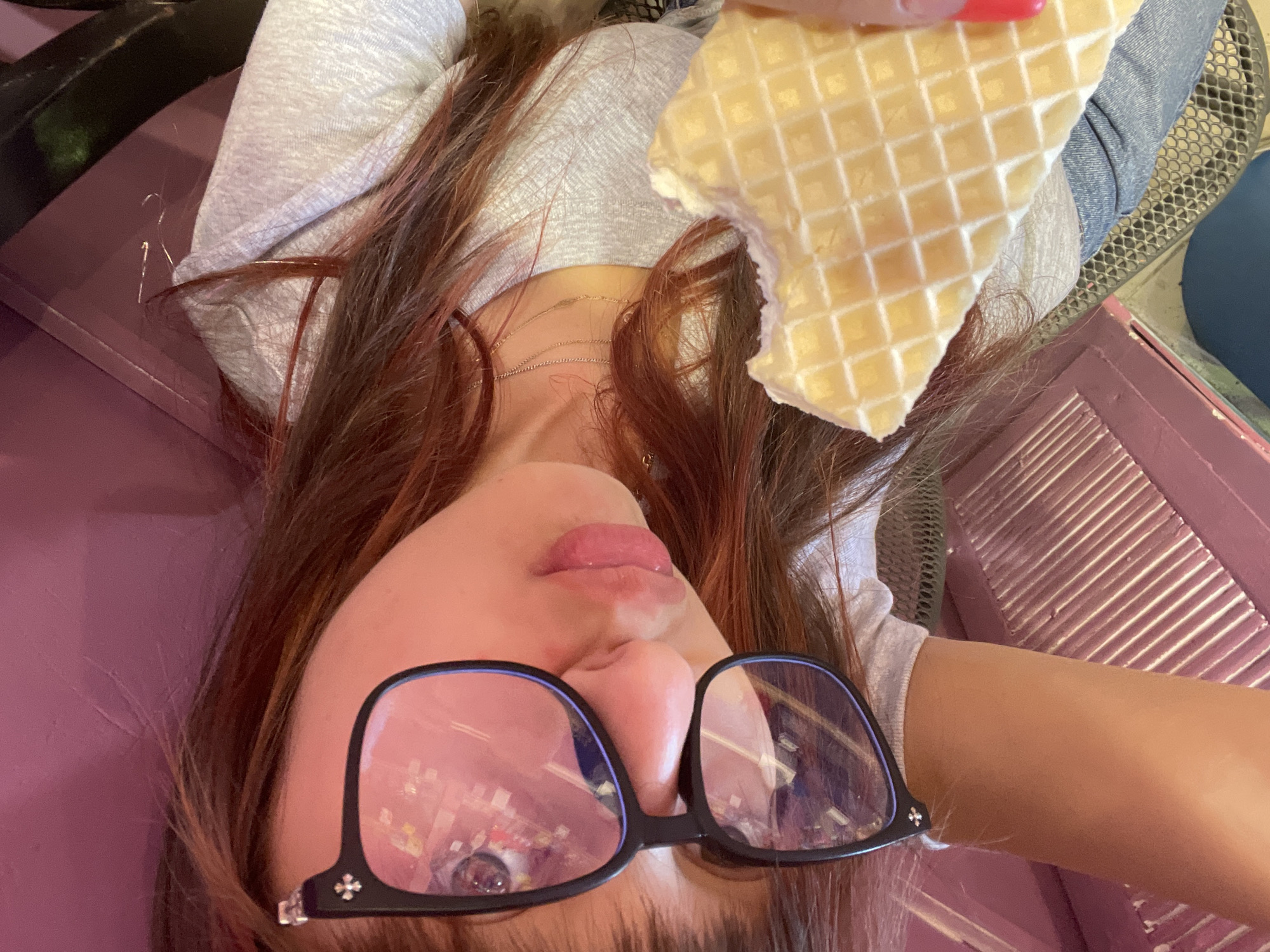 A young woman with long brown hair is taking a selfie while eating a waffle. She is wearing large black-rimmed glasses with a slight purple tint, a light gray t-shirt, and denim jeans. The reflection in her glasses shows lights and possibly a ceiling. Her left arm, visible in the lower left of the image, is resting on a pink surface. She is holding a bite out of a light-colored waffle with her right hand, which has bright red nail polish. The background is out of focus, but appears to be an indoor setting with pink and dark elements. There is no identifiable information about the specific location within Raanana, Israel.