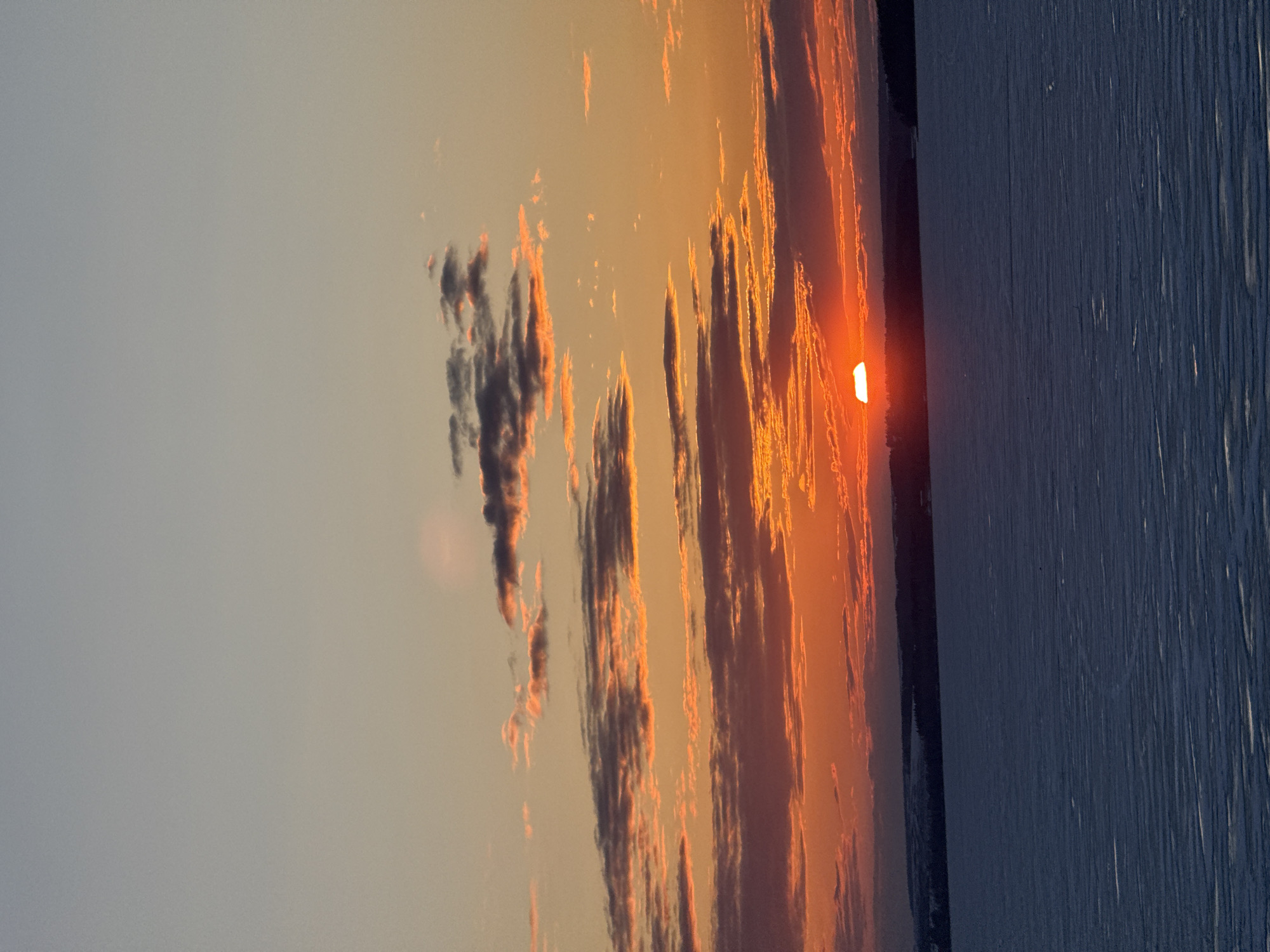 The image shows a sunset over what appears to be a body of water. The sun is a bright orange orb partially obscured by dark clouds that are vertically oriented, with orange highlighting. The sky above the clouds is a pale grey color. The water is textured with lines. A dark land mass separates the water from the sky. The location is possibly near or around Votkinsk, Russia, but there is no definitive visual evidence to confirm this within the image itself.