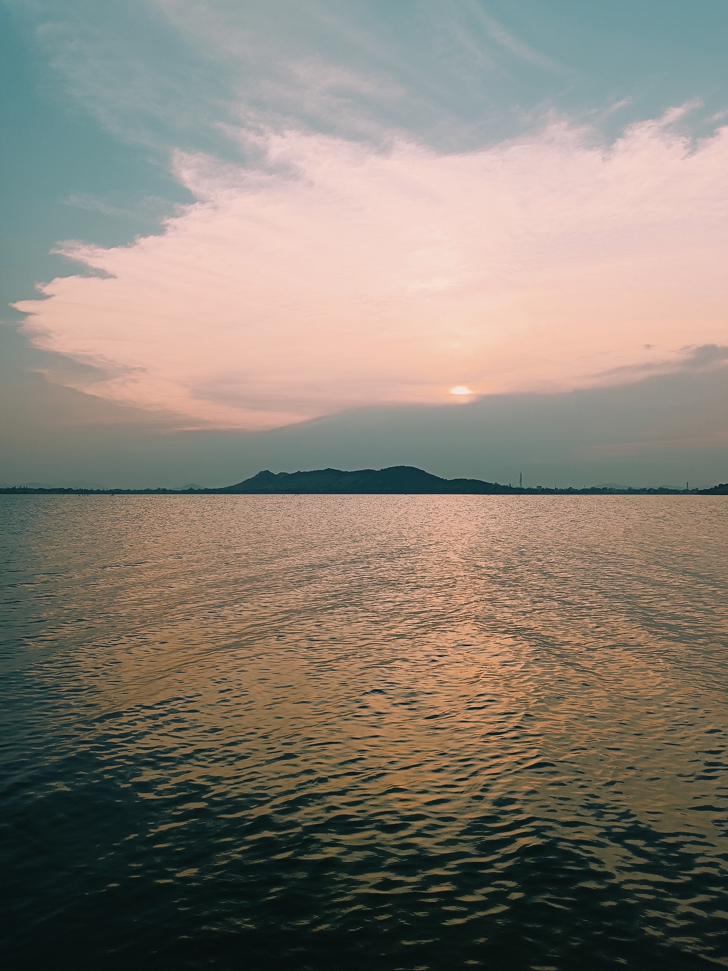 The image shows a wide expanse of water, likely a lake or large body of water, under a dusky sky. The sky transitions from a pale blue near the top to a soft pink and orange hue towards the horizon, where the sun is partially obscured by clouds. A long, low silhouette of a hill or small mountain stretches across the horizon line. On the right side, there are faint indications of man-made structures like transmission towers, which hint at a settlement or city nearby. The water's surface is rippled, reflecting the colors of the sky with variations in shades of orange and darker hues nearer to the viewer. The overall impression is serene and subdued. The city is Kammavaripalli, India.