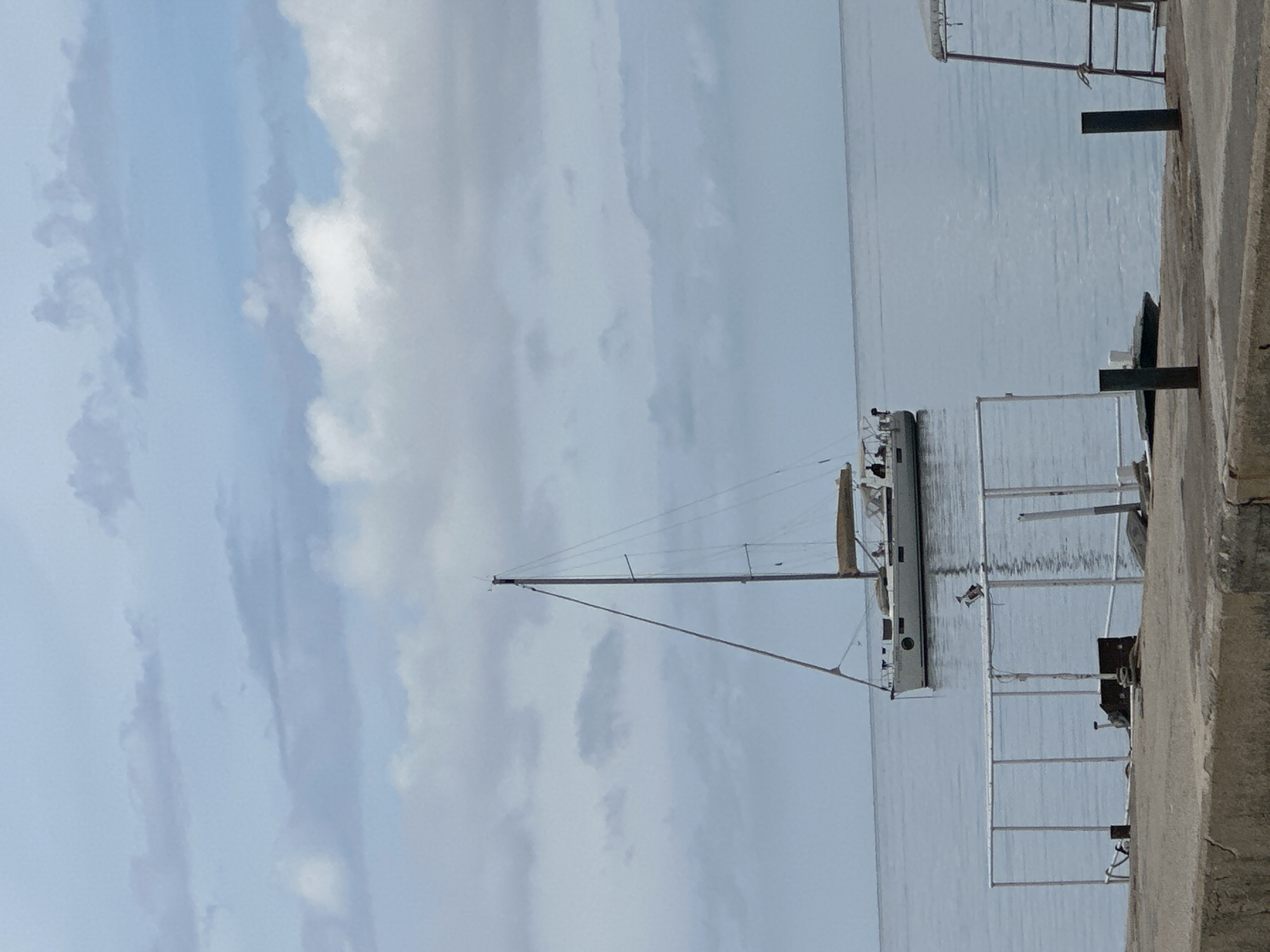The image shows a sailboat docked in the city of Budva, Montenegro. The boat is docked alongside a concrete pier. The sky is cloudy, and the water is calm.