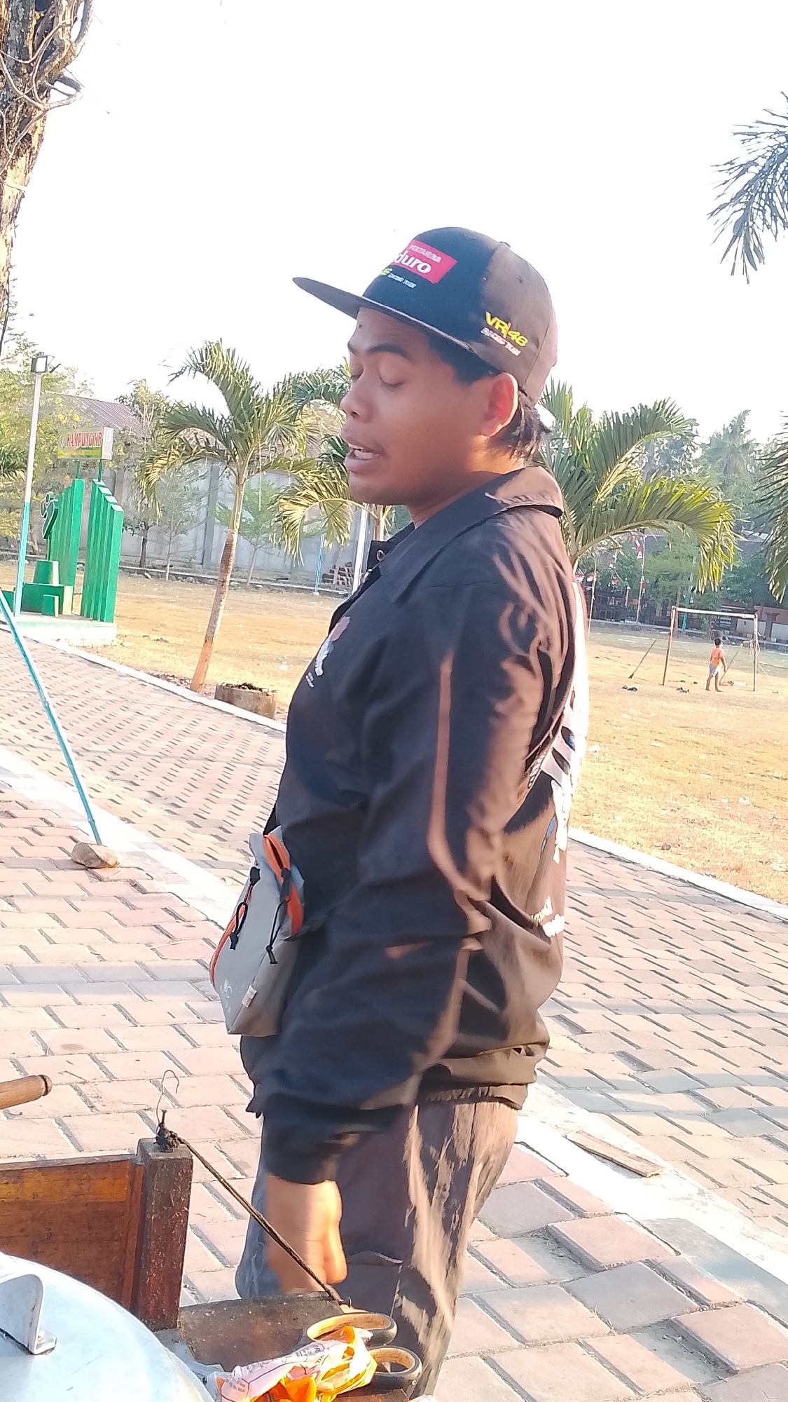 The image captures a medium shot of a young man standing outdoors, facing right in profile. He is wearing a black baseball cap with red and white lettering and a black collared jacket over a light-colored shirt. The jacket has a logo on the left chest and a larger design on the back. A small gray and orange bag is slung across his chest. His right hand is visible, holding a thin metal object. In the foreground, a wooden box with some metal objects and a pair of scissors is visible.
The background suggests a park or recreational area. Palm trees and other foliage are present, along with a paved pathway made of brick-like material. In the distance, a soccer goal and two small figures, one in an orange shirt, are visible on a grassy field. There is also a green structure in the middle left of the image, possibly part of a playground or park amenity. The lighting indicates it is daytime, likely late afternoon due to the long shadows and warm light. The overall setting appears to be outdoors in a tropical or subtropical climate, consistent with a location like Karanglangu, Indonesia.